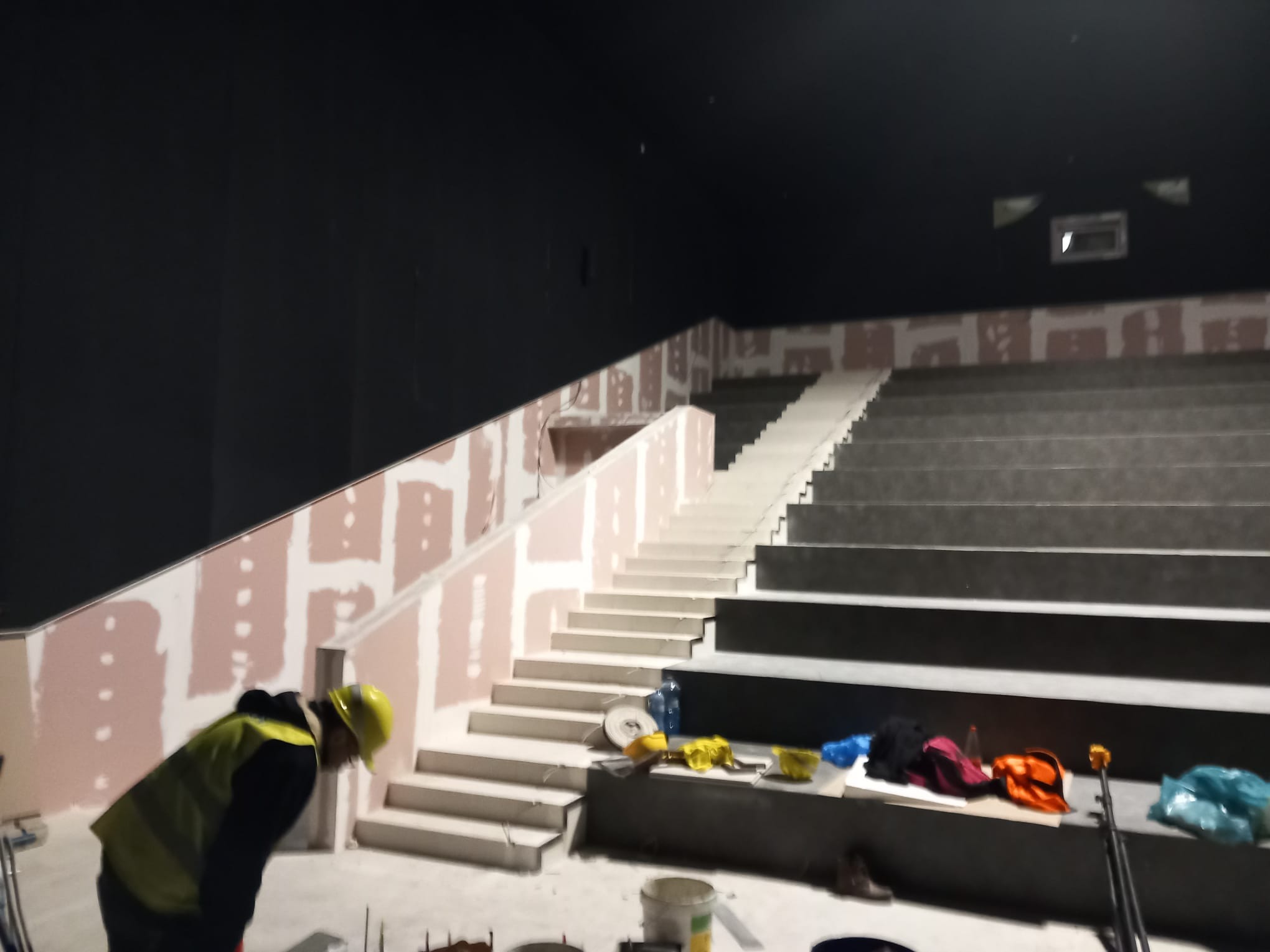 Montaż stopni schodów w sali kinowej, widoczny pracownik w żółtej kamizelce, narzędzia i materiały budowlane na schodach, ściany przygotowane do malowania.