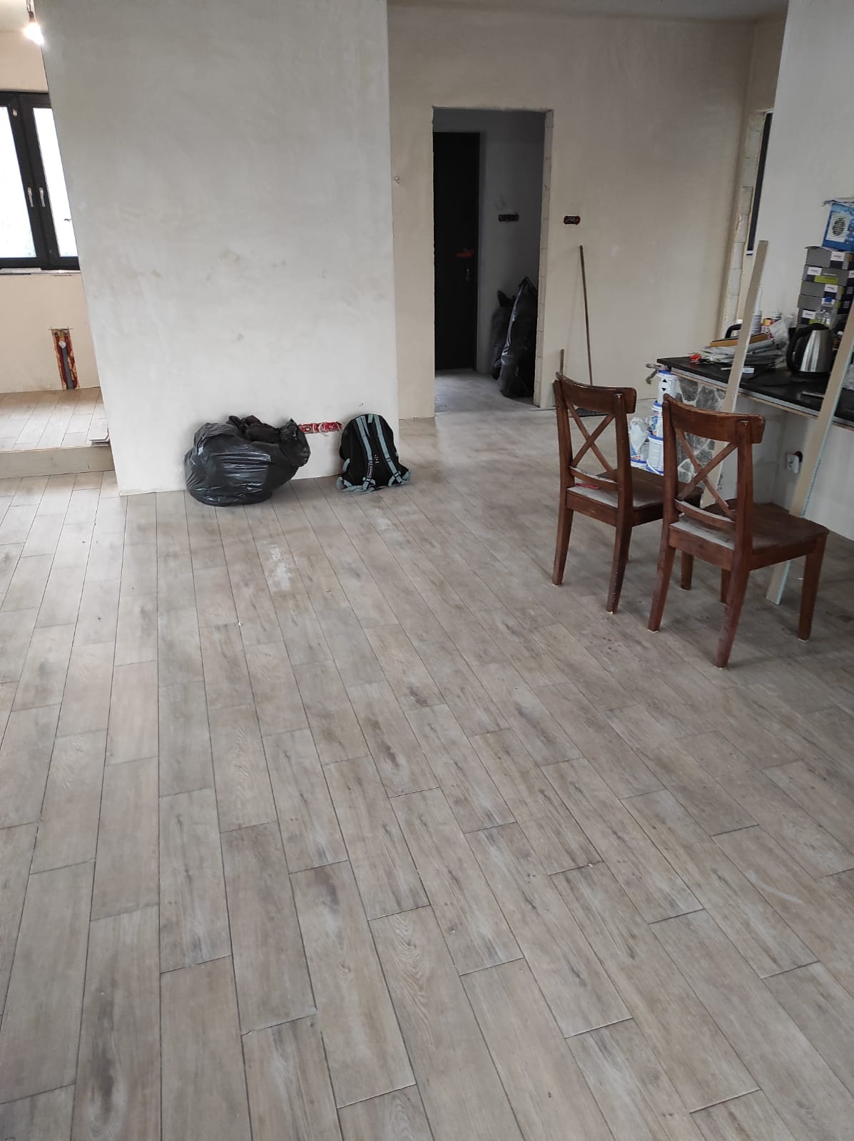 Jasne, podłużne płytki imitujące drewno, ułożone na podłodze w trakcie remontu, widoczne dwa krzesła, worki z odpadami budowlanymi oraz plecak robotnika.