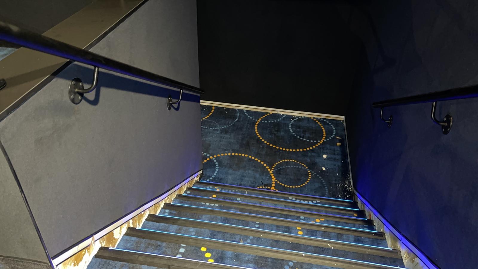 Schody pokryte wzorzystą wykładziną z podświetleniem LED, widok z góry, ciemne ściany i czarne poręcze.