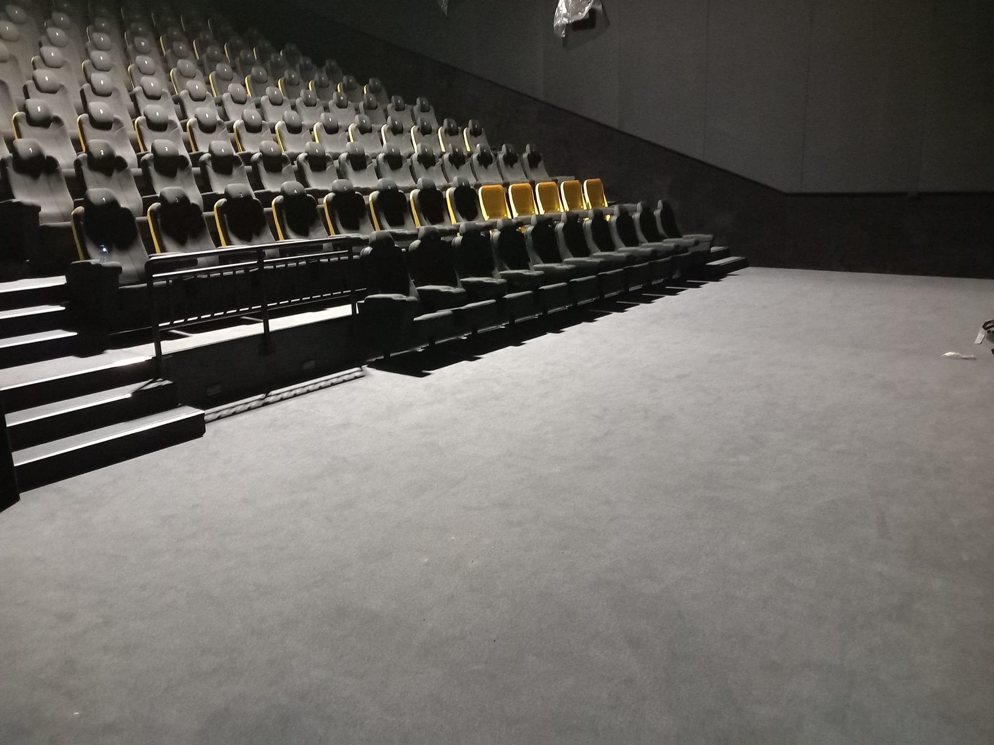 Sala kinowa z rzędami czarnych foteli z żółtymi akcentami, pokryta szarą wykładziną dywanową.