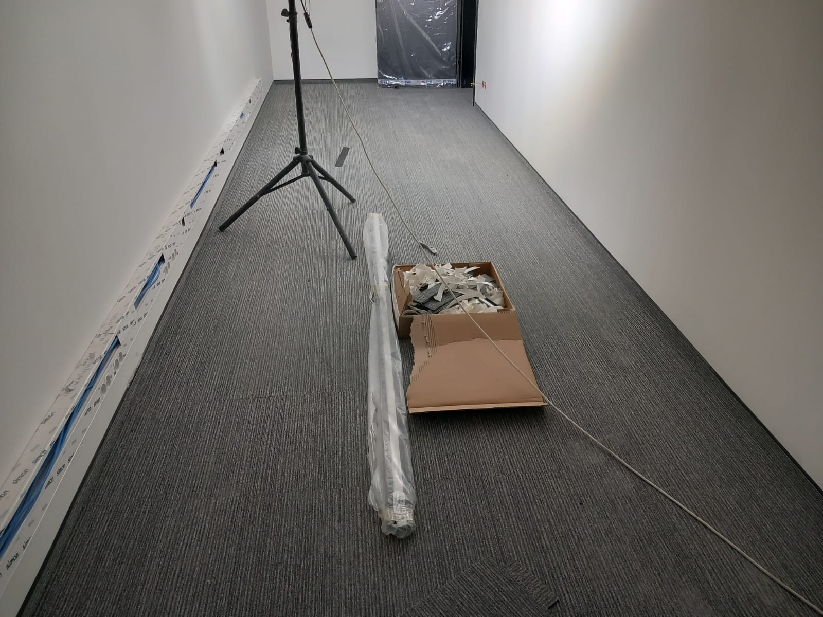 Korytarz z szarą wykładziną dywanową w trakcie instalacji, widoczne listwy przypodłogowe w opakowaniach, karton z odpadami i statyw oświetleniowy.