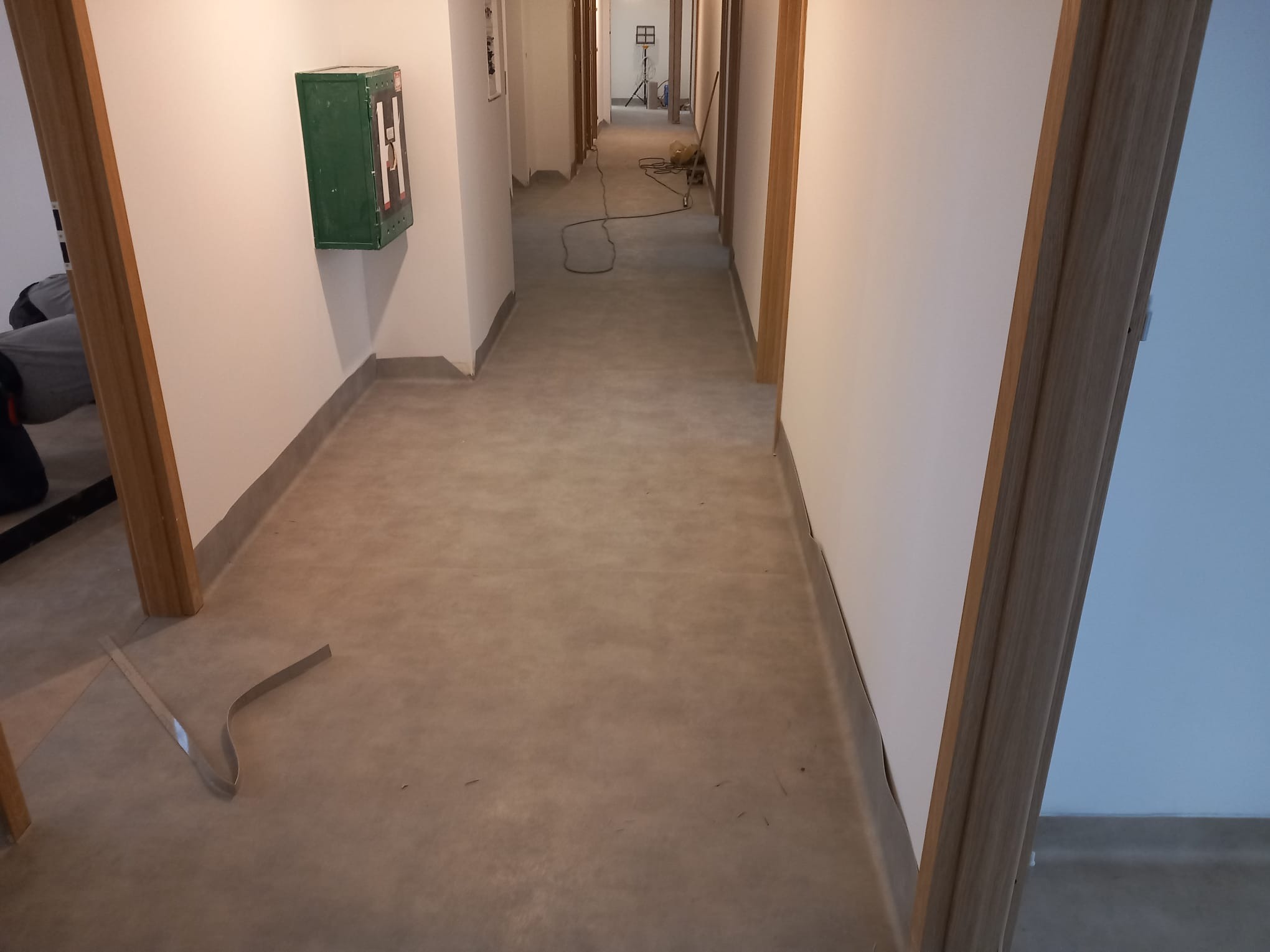 Korytarz z nową, jednolitą wykładziną podłogową w odcieniach szarości, widoczna listwa przypodłogowa i fragmenty narzędzi przy progu.