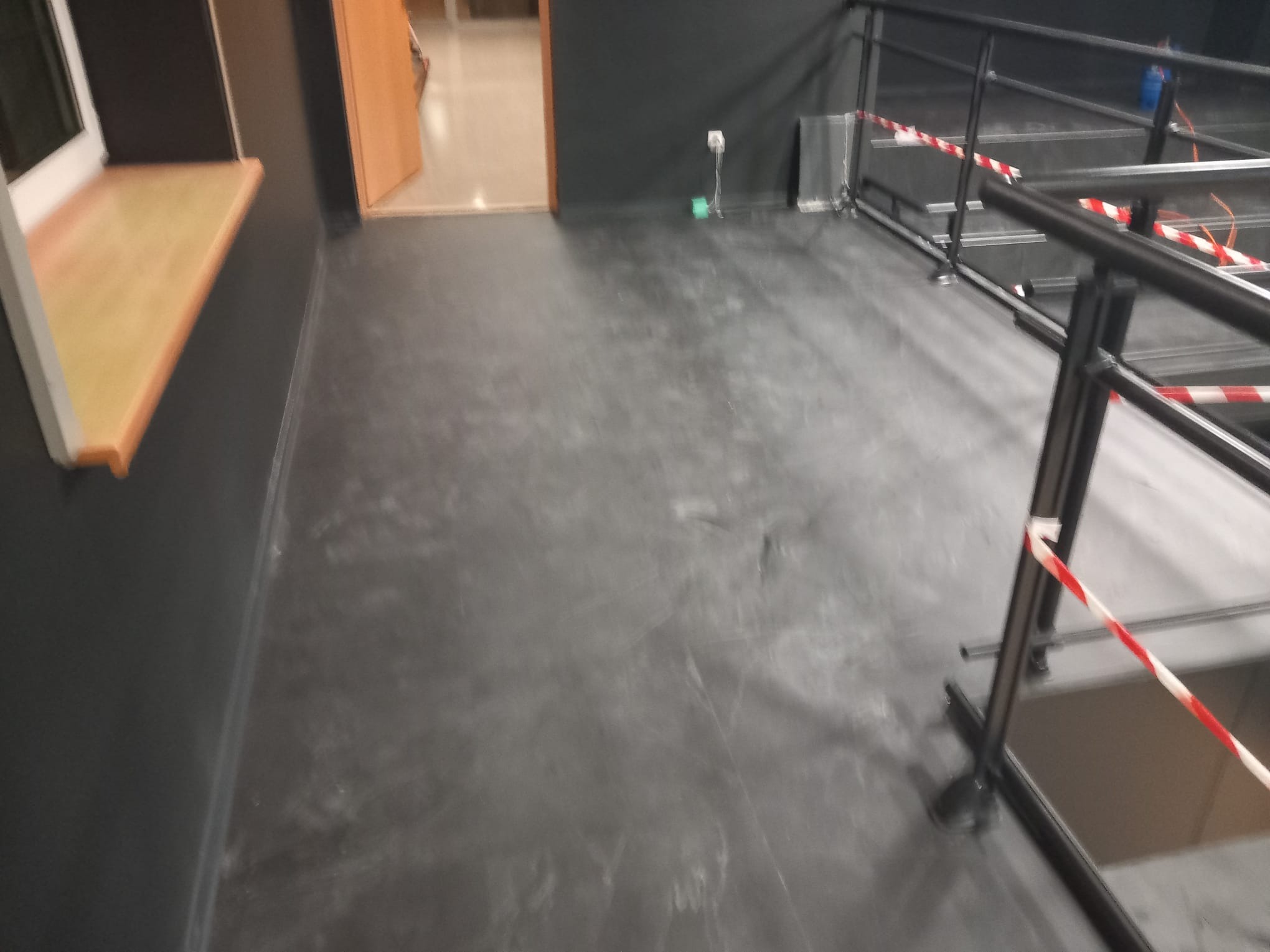 Szara, gładka podłoga w pomieszczeniu z czarnymi ścianami, widoczne fragmenty okna i schodów z barierką zabezpieczoną taśmą ostrzegawczą.