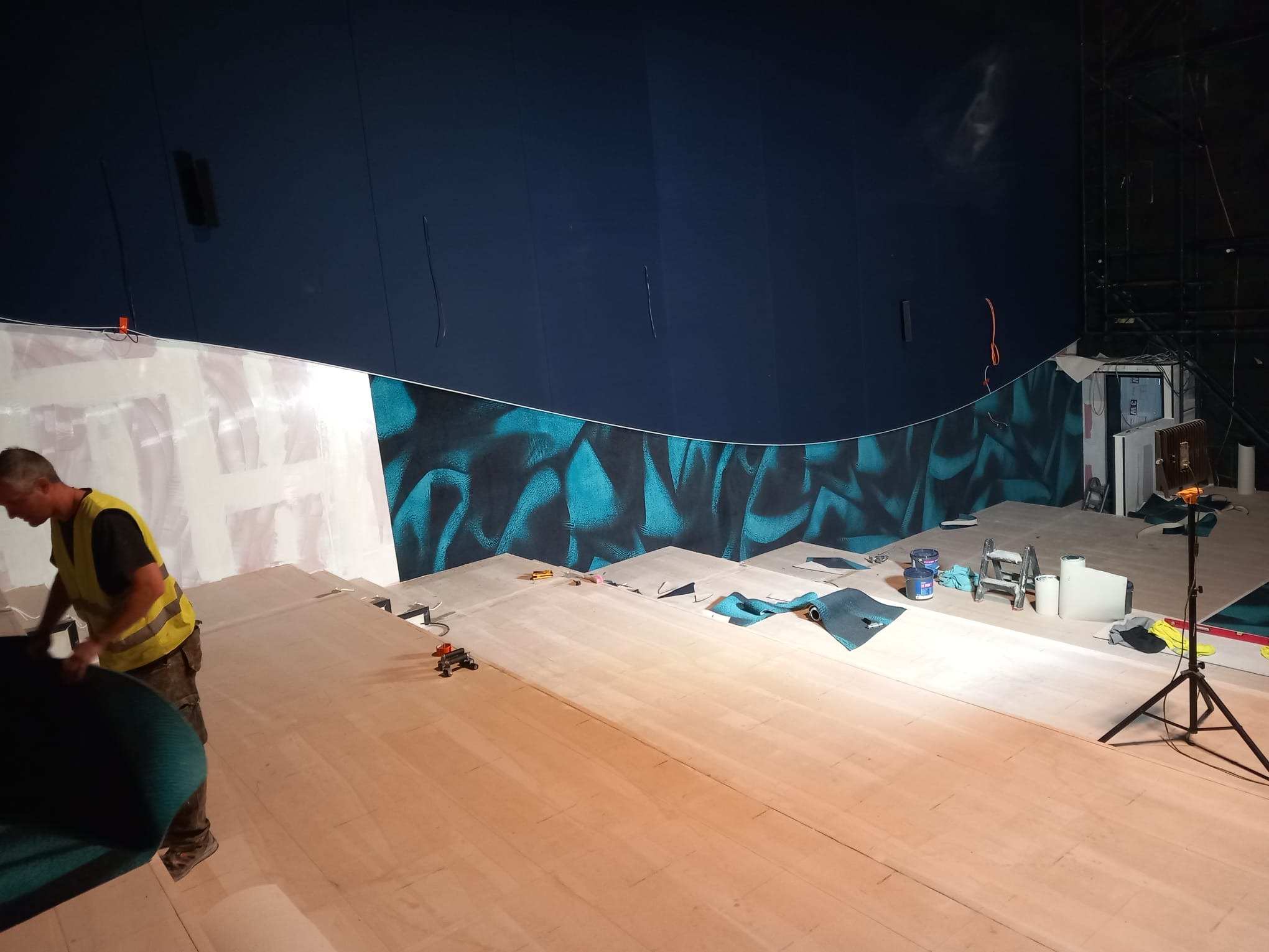 Montaż wykładziny podłogowej w nietypowym pomieszczeniu: pracownik w kamizelce odblaskowej układa zieloną wykładzinę na drewnianej podłodze na tle ciemnoniebieskiej ściany z abstrakcyjnym wzorem...