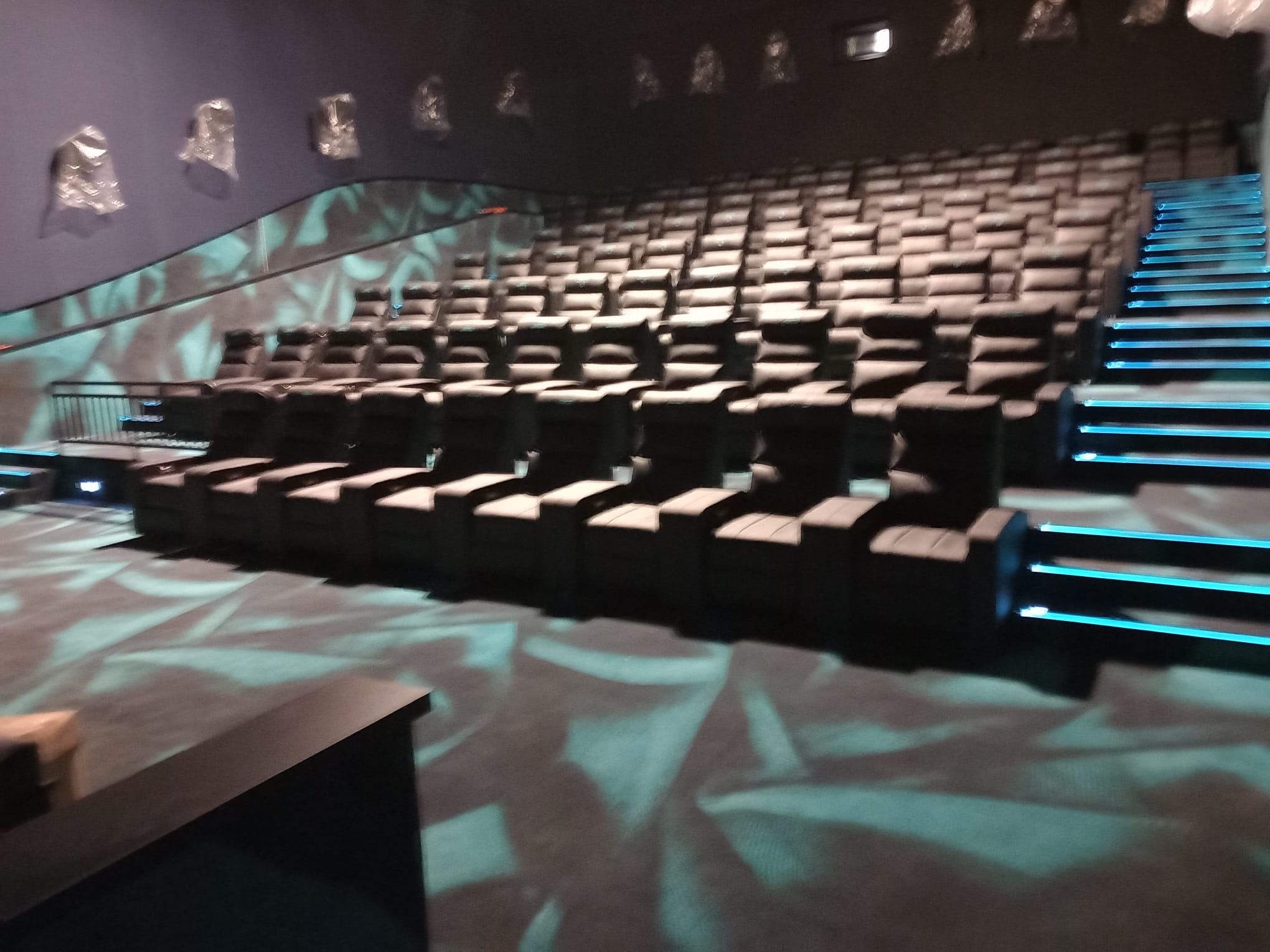 Rzędy ciemnych foteli w kinie, oświetlone schody z niebieskim podświetleniem, wzorzysta wykładzina podłogowa.