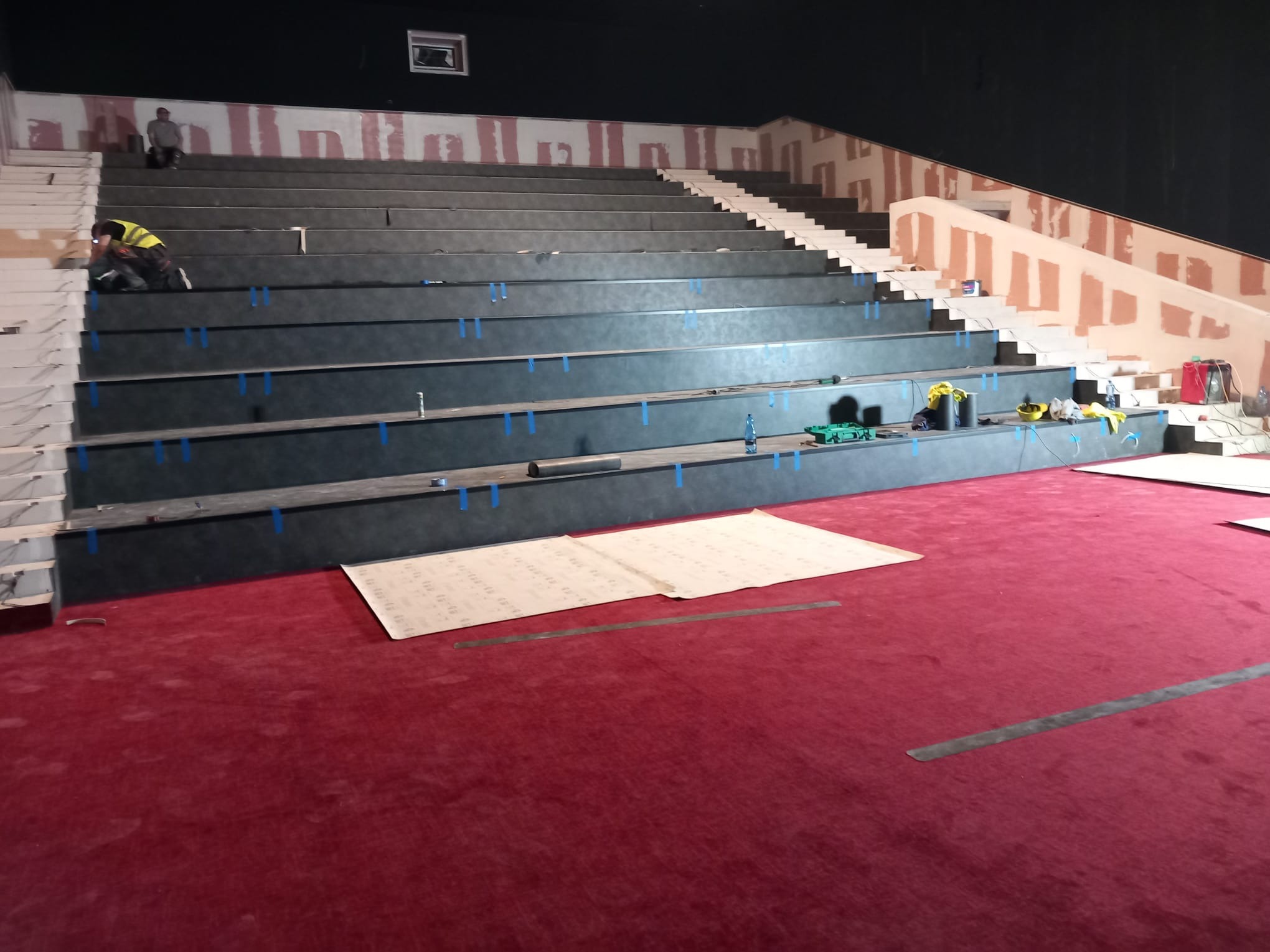 Montaż stopni schodów w kinie lub teatrze, widoczny pracownik w kamizelce odblaskowej, czerwona wykładzina na podłodze, etap prac wykończeniowych na ścianach.