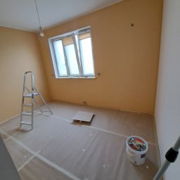 HDM - Pokój w trakcie odświeżania: ściany pomalowane na beżowo, zabezpieczona podłoga, drabina aluminiowa, wiadro z farbą i rękawicami na pokrywie.