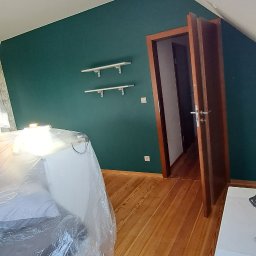 HDM - Pokój na poddaszu z drewnianą podłogą i ścianą pomalowaną na butelkową zieleń; meble zabezpieczone folią malarską, widoczne dwie białe półki na ścianie.