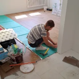 Pracownik w trakcie układania paneli podłogowych na zielonej macie wygłuszającej, widoczne narzędzia pomiarowe, częściowo ułożona podłoga i nieuprzątnięte pozostałości po pracach.