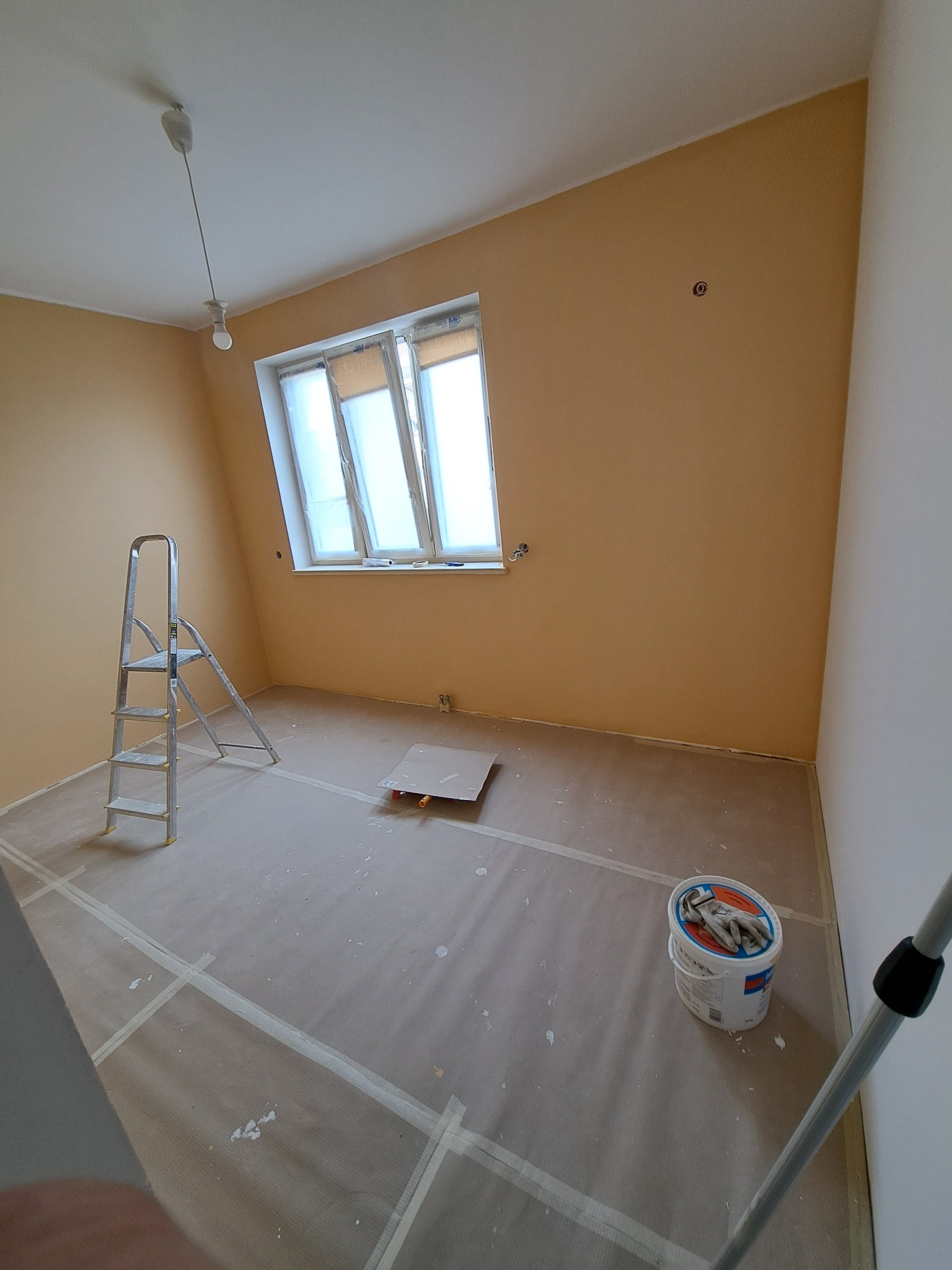 Pokój w trakcie odświeżania: ściany pomalowane na beżowo, zabezpieczona podłoga, drabina aluminiowa, wiadro z farbą i rękawicami na pokrywie.