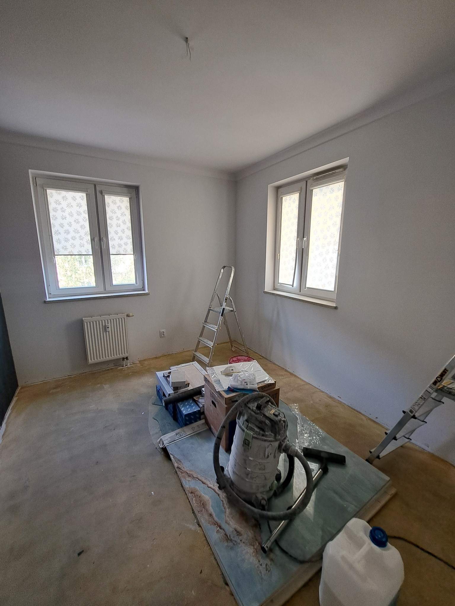 Pokój w trakcie remontu, ściany świeżo pomalowane na szaro, widoczne okna z roletami, drabina, odkurzacz przemysłowy i inne narzędzia malarskie.