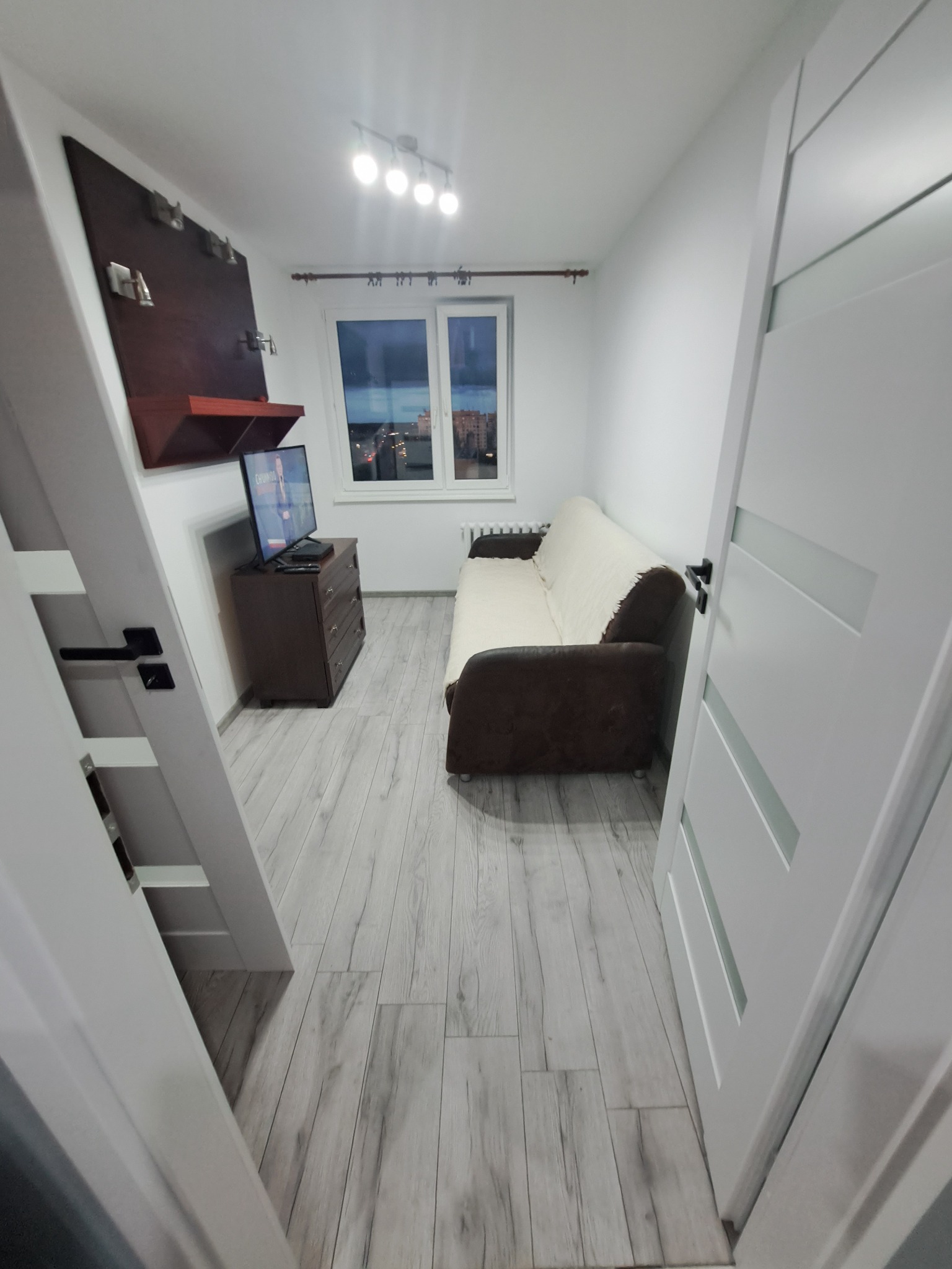 Wnętrze pokoju z szarymi panelami podłogowymi, brązową sofą z beżową narzutą, komodą z telewizorem i białą ścianą z brązową półką, widoczne przez otwarte białe drzwi z przeszkleniami.