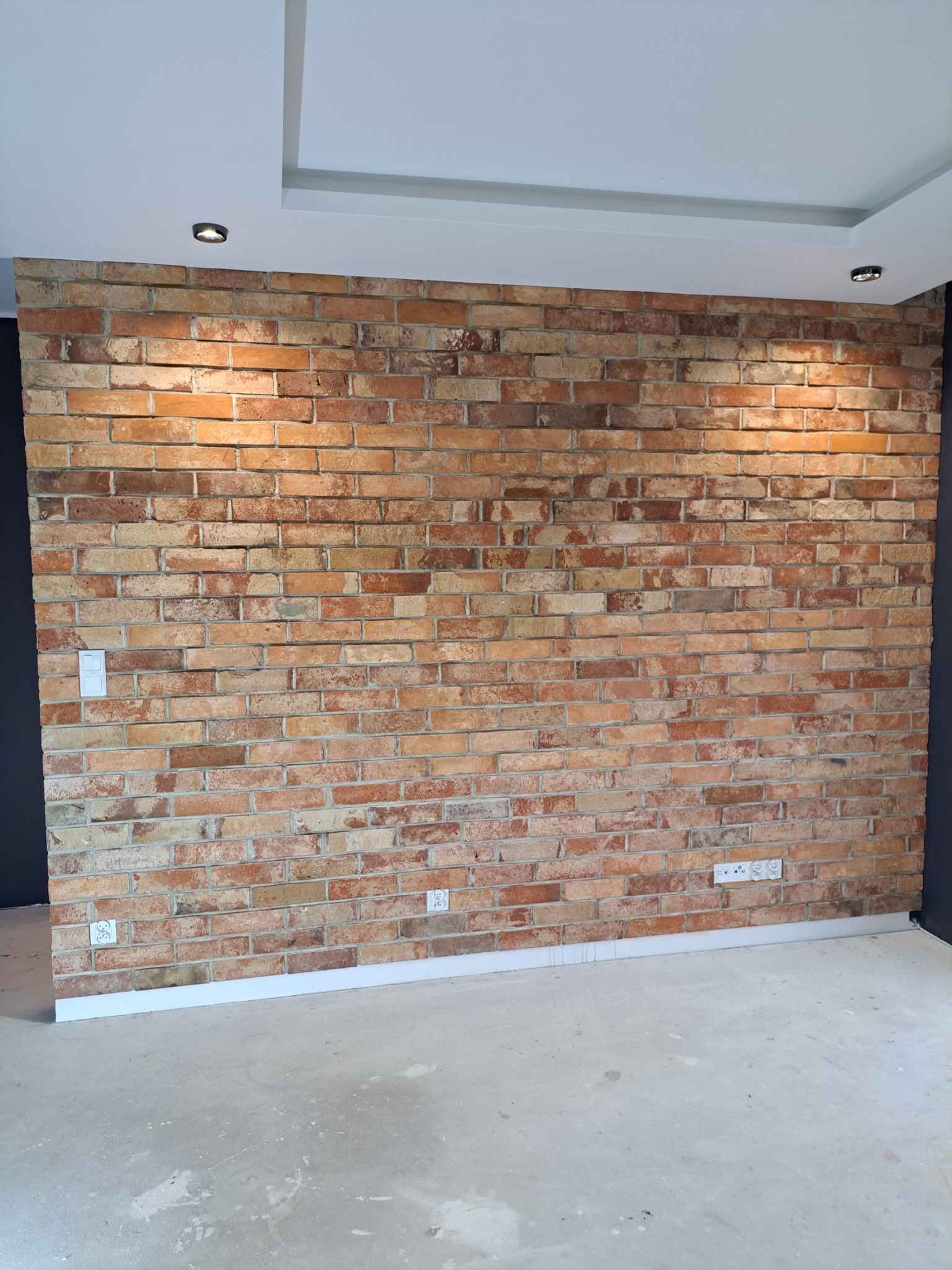 Ściana z cegły klinkierowej w nowym wnętrzu, oświetlona punktowymi lampami sufitowymi, z widocznymi gniazdkami elektrycznymi i białymi listwami przypodłogowymi na tle szarej ściany i surowej podłogi.