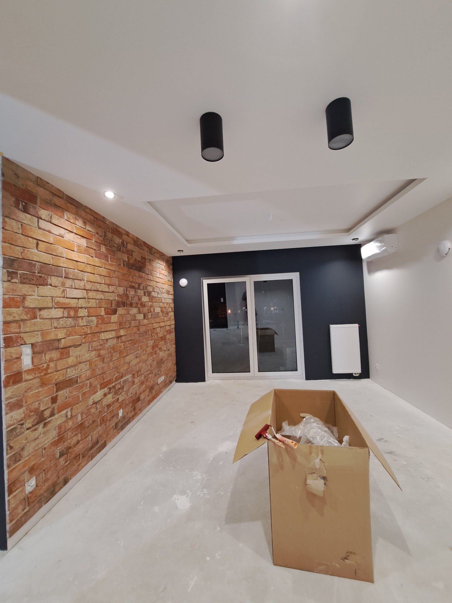 Pokój po remoncie z ceglaną ścianą, czarną ścianą z oknem balkonowym, białym grzejnikiem i kartonem z narzędziami malarskimi na podłodze.