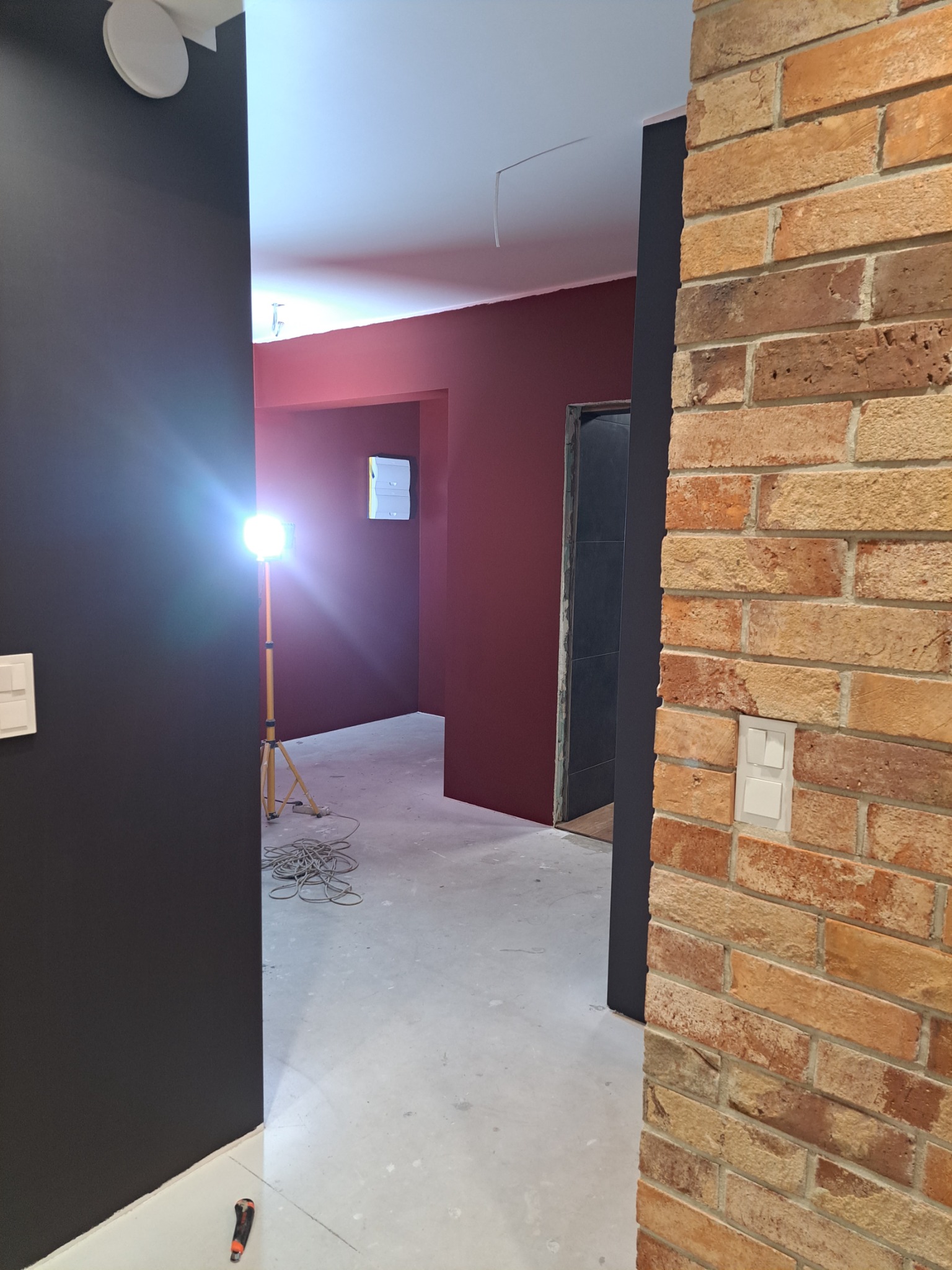 Wnętrze w trakcie remontu: pomalowane na bordowo ściany, surowa podłoga, ceglana ściana z włącznikiem, lampa budowlana oświetlająca pomieszczenie, widoczne przewody i narzędzie na podłodze.
