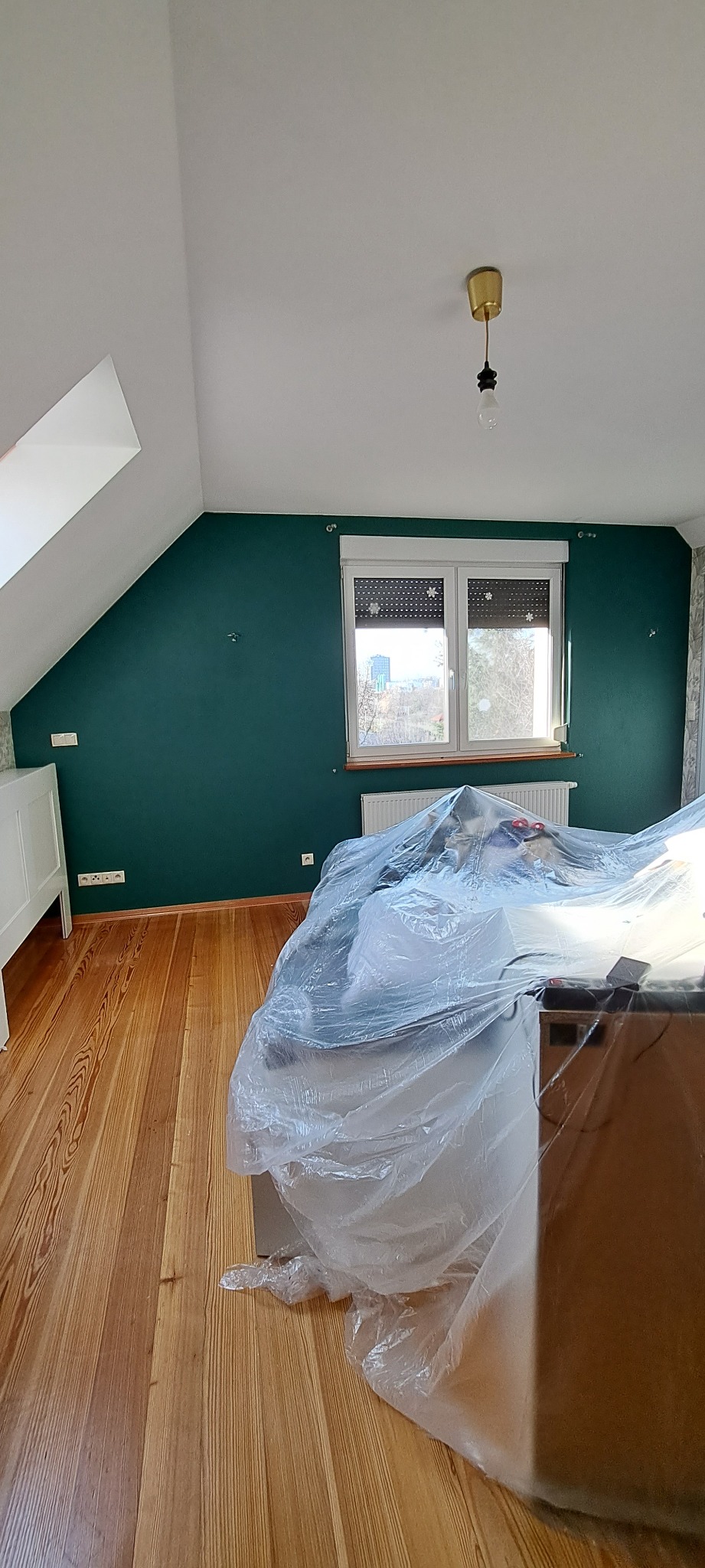 Pokój na poddaszu z drewnianą podłogą, ściana pomalowana na ciemnozielono, okno z roletami, meble zabezpieczone folią malarską, przygotowania do prac wykończeniowych.