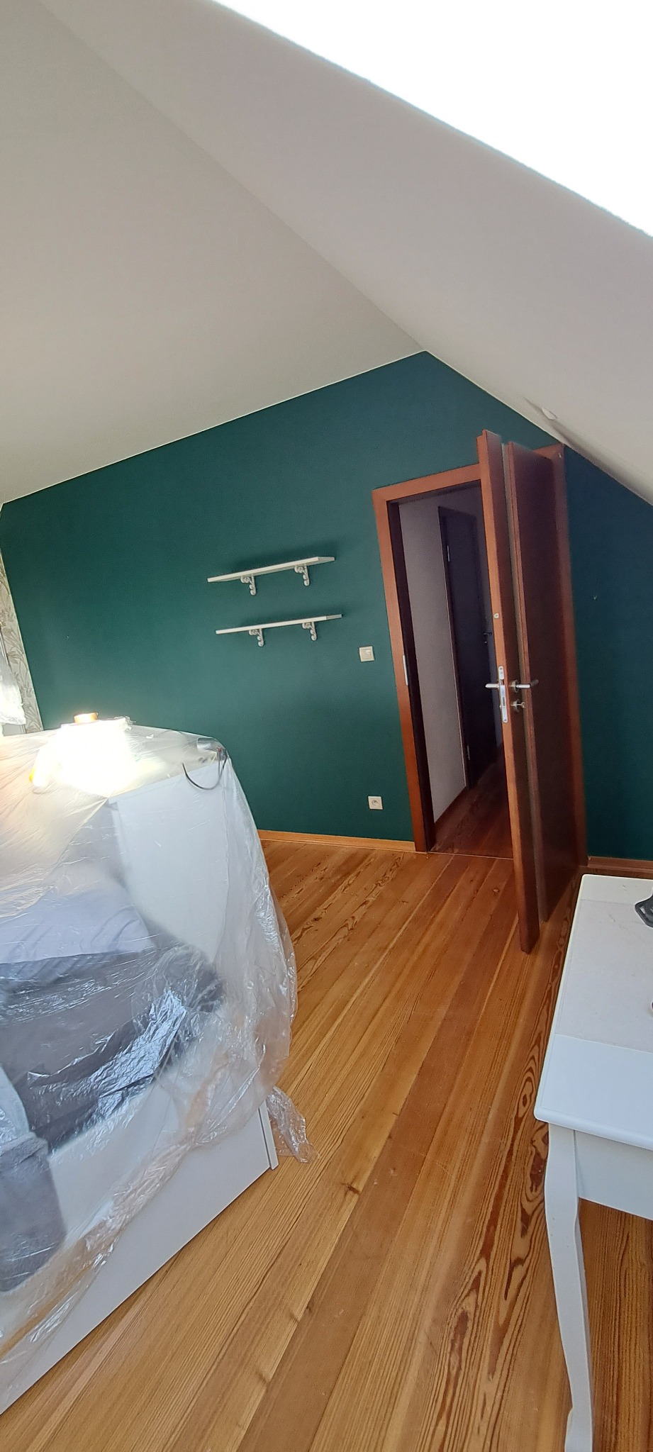 Pokój na poddaszu z drewnianą podłogą i ścianą pomalowaną na butelkową zieleń; meble zabezpieczone folią malarską, widoczne dwie białe półki na ścianie.