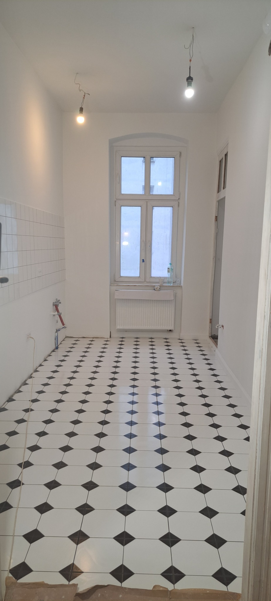 Wnętrze kuchni po remoncie, świeżo ułożona podłoga z białych płytek z czarnymi rombami, białe ściany z fragmentem glazury, okno z widokiem na zewnątrz, surowa żarówka zwisająca z sufitu.