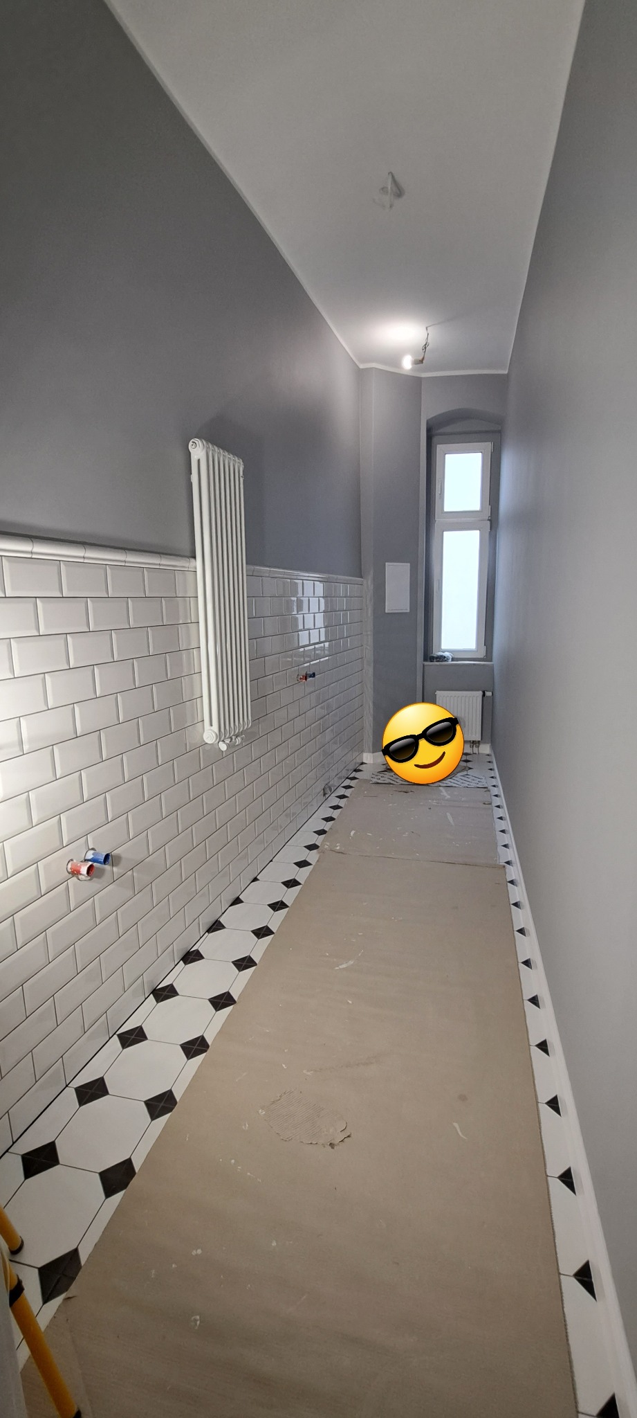 Wąska łazienka w trakcie remontu: białe, prostokątne płytki na ścianach, szare ściany powyżej, podłoga zabezpieczona kartonem, widoczne przyłącza hydrauliczne i grzejnik.