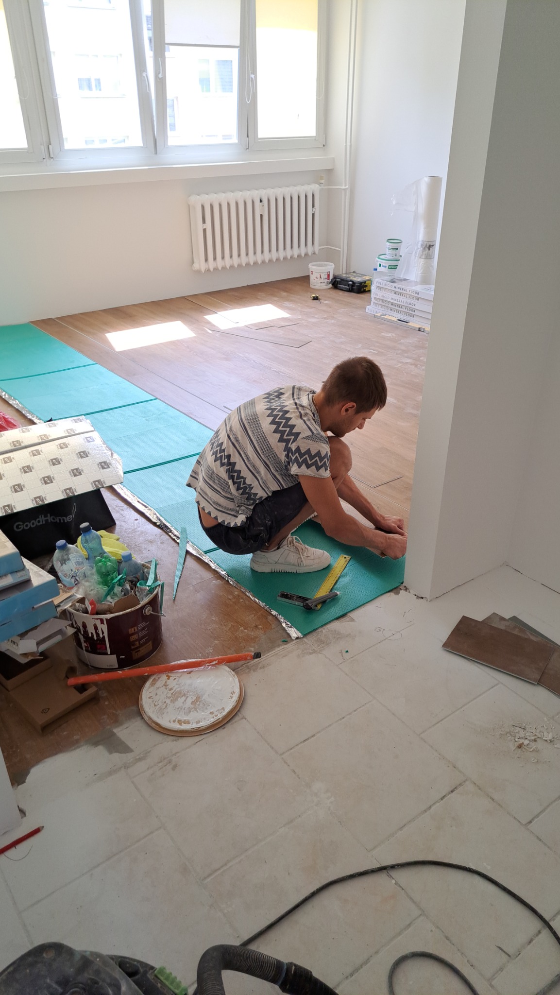 Pracownik w trakcie układania paneli podłogowych na zielonej macie wygłuszającej, widoczne narzędzia pomiarowe, częściowo ułożona podłoga i nieuprzątnięte pozostałości po pracach.