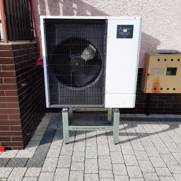 Opal-House - Biała zewnętrzna jednostka pompy ciepła Mitsubishi Zubadan R32, zamontowana na metalowej podstawie na kostce brukowej, z widoczną obudową licznika elektrycznego.