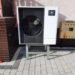 Biała zewnętrzna jednostka pompy ciepła Mitsubishi Zubadan R32, zamontowana na metalowej podstawie na kostce brukowej, z widoczną obudową licznika elektrycznego.