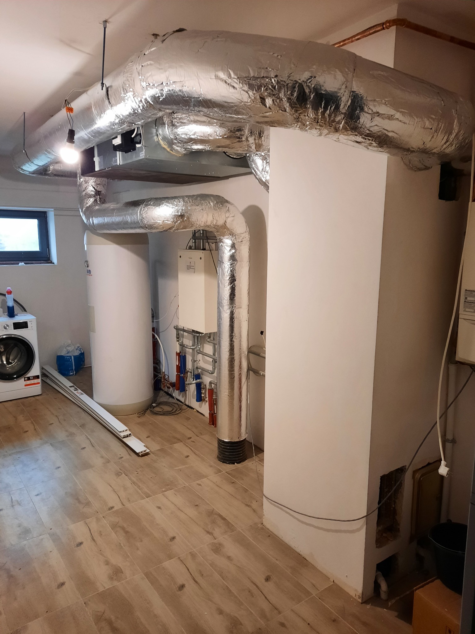 Kotłownia z piecem, zasobnikiem ciepłej wody i instalacją grzewczą z izolowanymi rurami, pralka w tle.