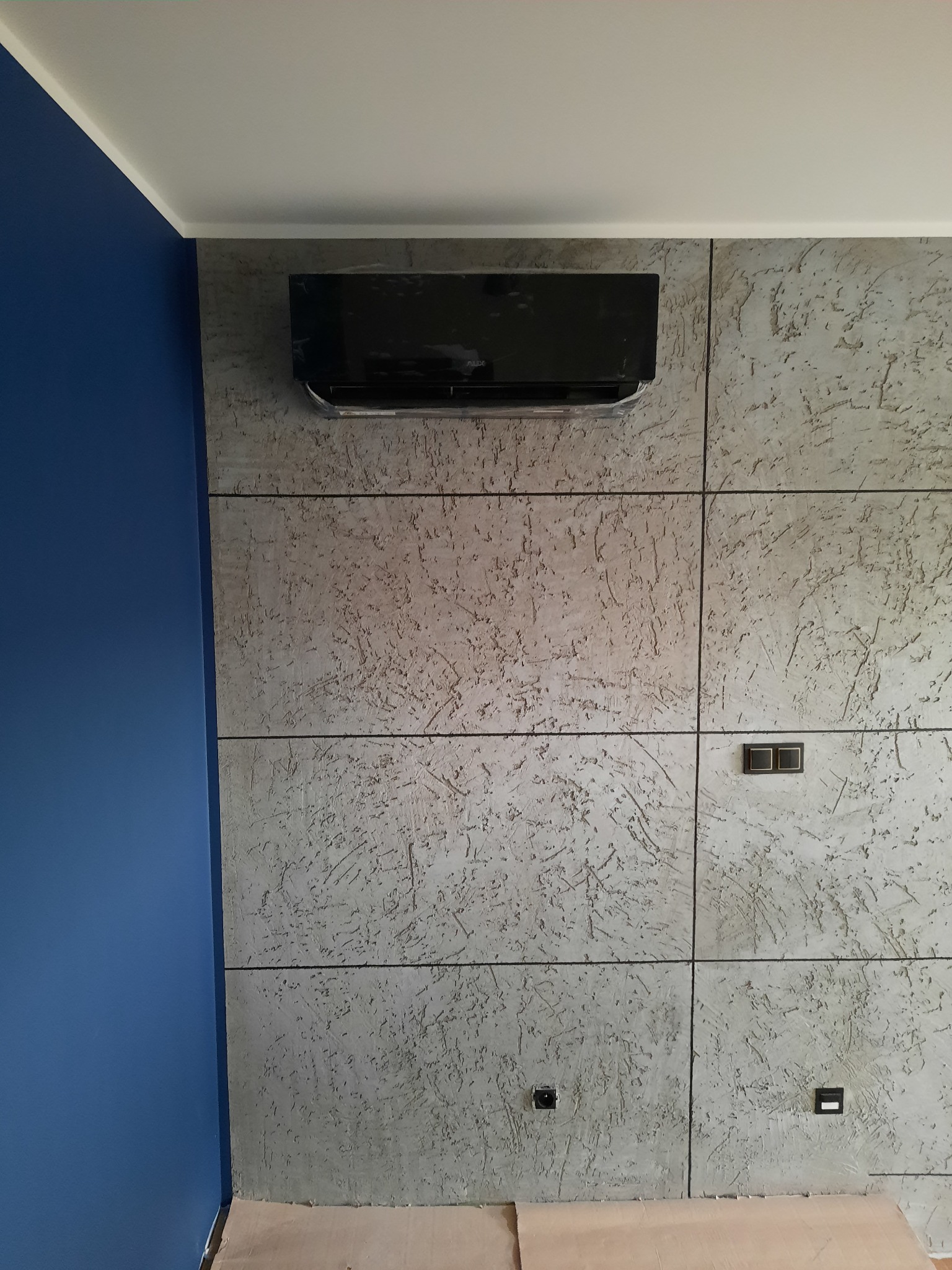 Czarny klimatyzator ścienny zamontowany na szarej ścianie z fakturą, widoczne gniazdka elektryczne i fragment niebieskiej ściany obok.