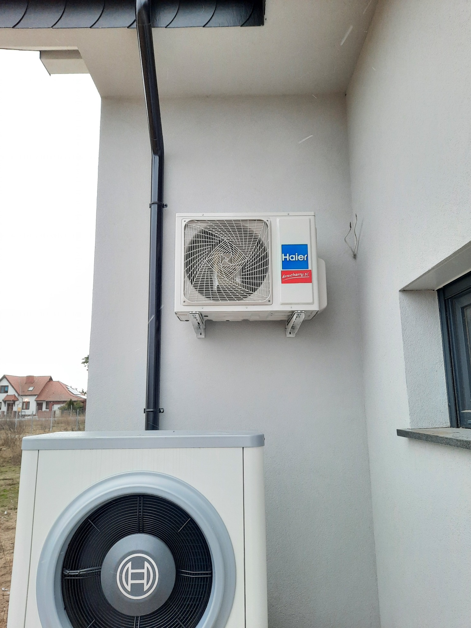 Zewnętrzna jednostka klimatyzatora Haier zamontowana na ścianie budynku obok rury spustowej i okna, z dolną jednostką pompy ciepła Bosch widoczną poniżej.