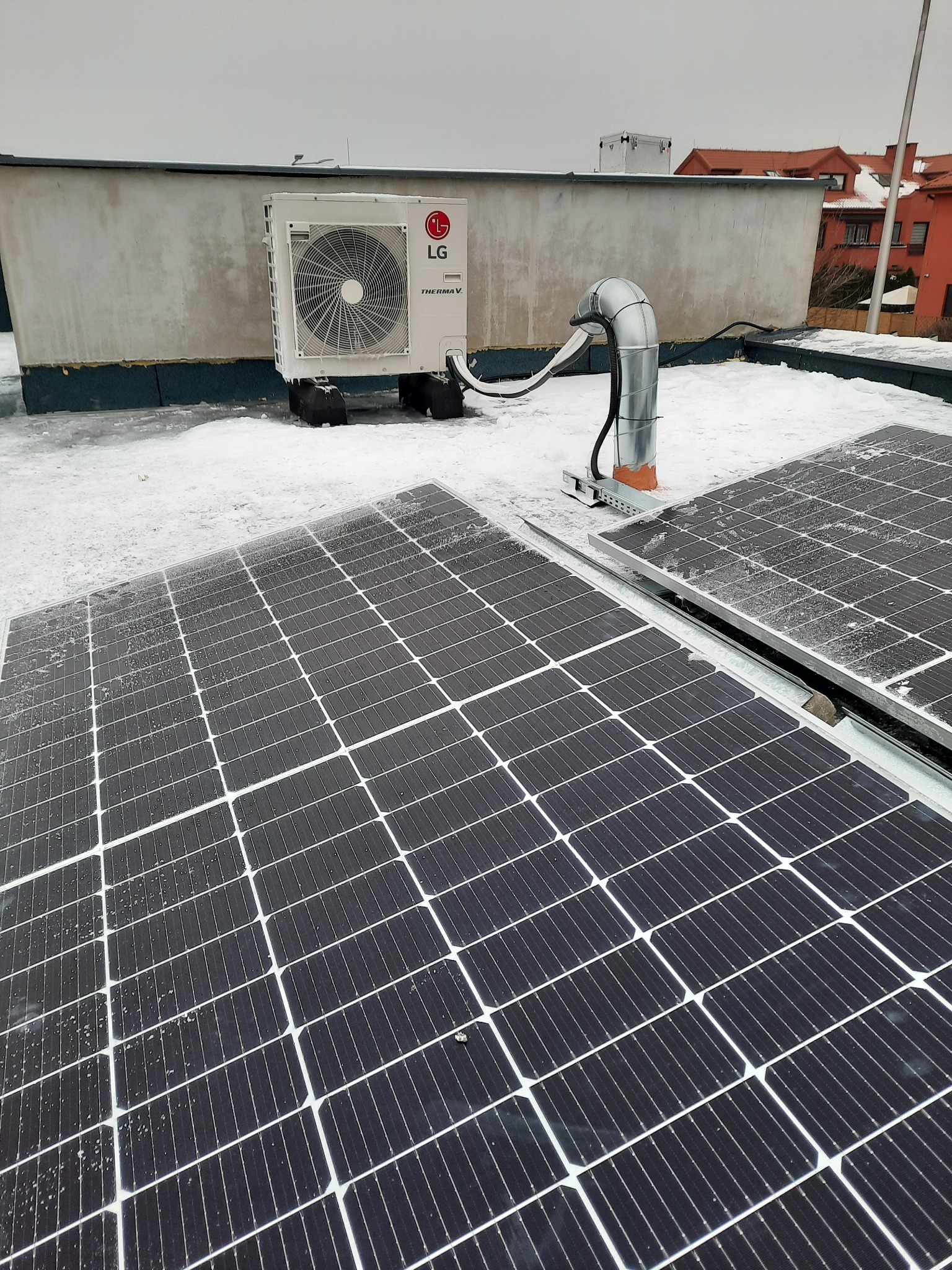 Instalacja paneli słonecznych i jednostki zewnętrznej klimatyzacji LG Therma V na płaskim, ośnieżonym dachu budynku.