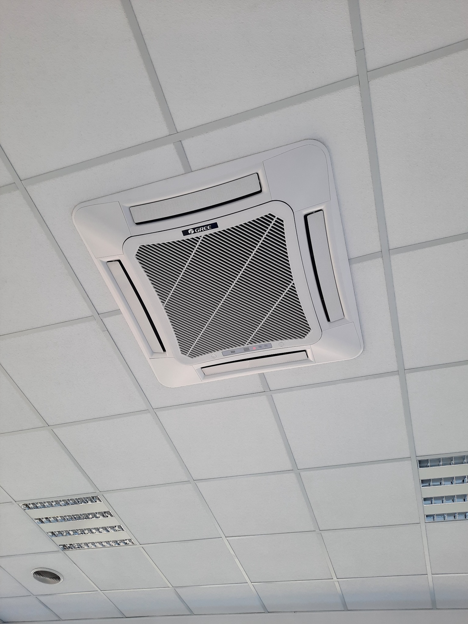 Biała kasetonowa jednostka klimatyzacyjna marki Gree zamontowana w suficie podwieszanym z widocznymi kratkami wentylacyjnymi i oświetleniem rastrowym w tle.