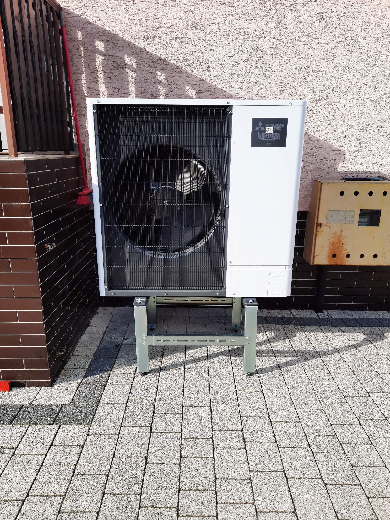 Biała zewnętrzna jednostka pompy ciepła Mitsubishi Zubadan R32, zamontowana na metalowej podstawie na kostce brukowej, z widoczną obudową licznika elektrycznego.