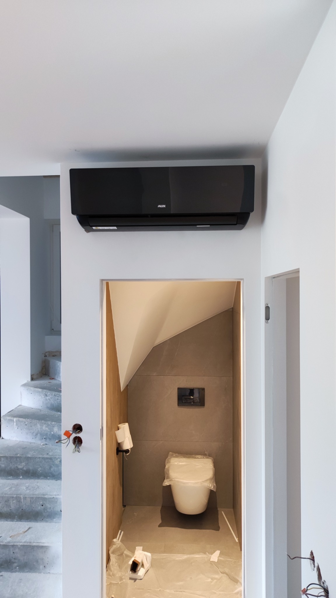 Czarny klimatyzator AUX zamontowany nad wejściem do łazienki w trakcie remontu, widoczne niedokończone schody i zabezpieczona folią toaleta.