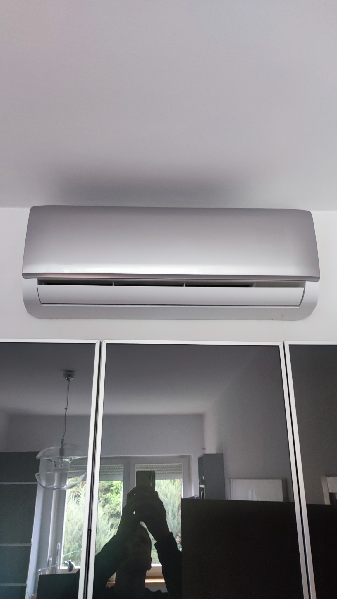Srebrny klimatyzator ścienny zamontowany w pomieszczeniu, widoczne odbicie osoby robiącej zdjęcie w szklanej powierzchni szafy.