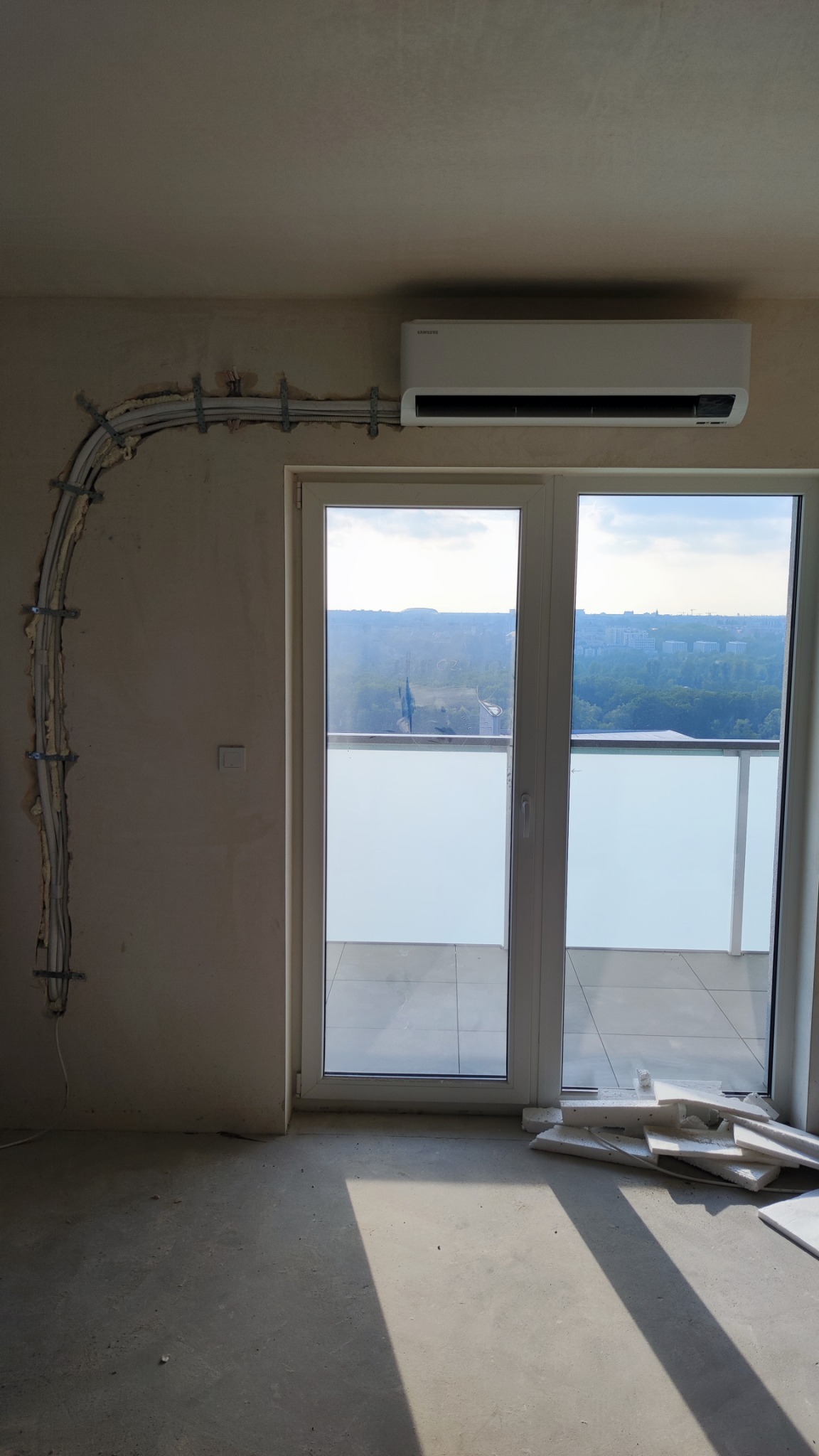 Montaż klimatyzatora Samsung w surowym wnętrzu, widoczne kable w korytku przymocowanym do ściany, okno balkonowe z widokiem na miasto.