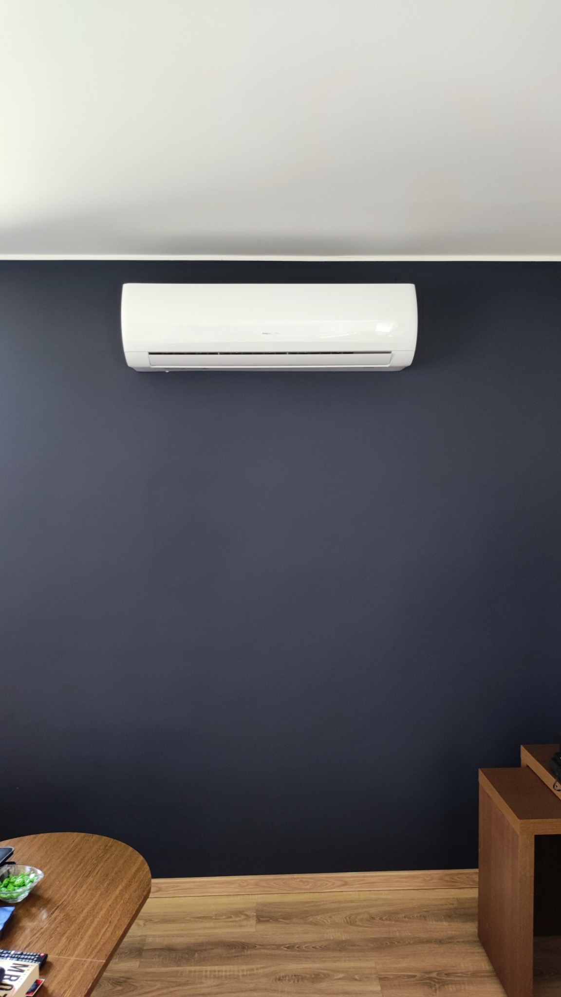 Biała jednostka wewnętrzna klimatyzacji zamontowana na ciemnogranatowej ścianie w nowoczesnym wnętrzu, widoczny fragment drewnianego stolika i podłogi.