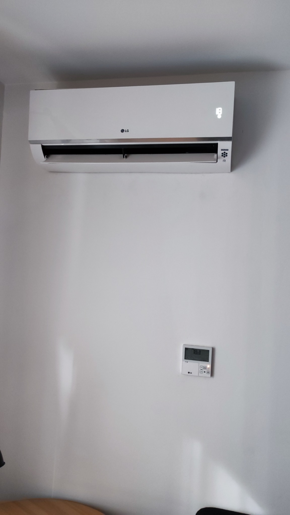 Biały klimatyzator LG zamontowany na białej ścianie, z widocznym panelem sterowania pokazującym temperaturę 18 stopni Celsjusza.