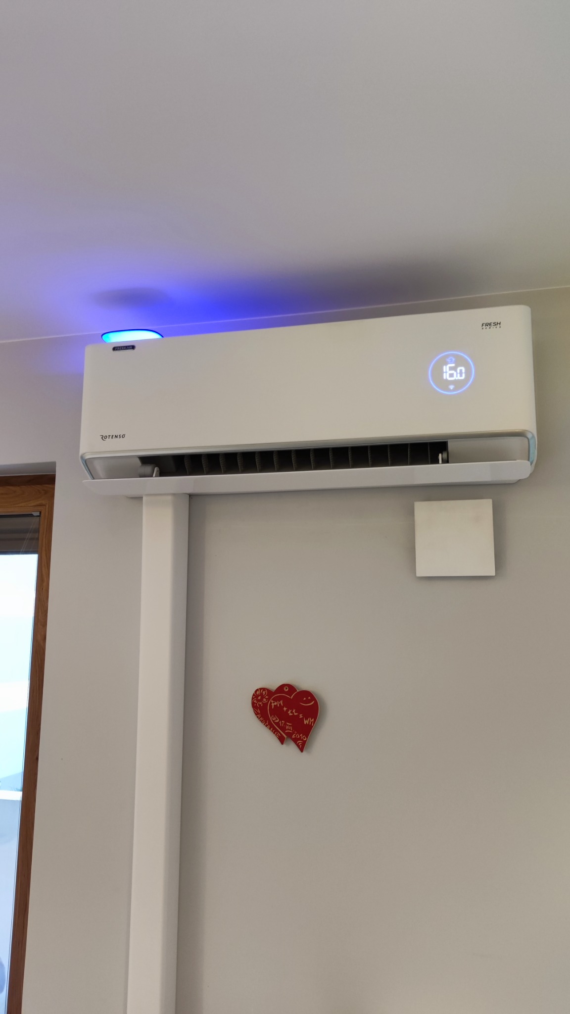 Biała klimatyzacja Rotenso FRESH z niebieską poświatą LED na ścianie, pod nią dekoracja w kształcie dwóch czerwonych serc.