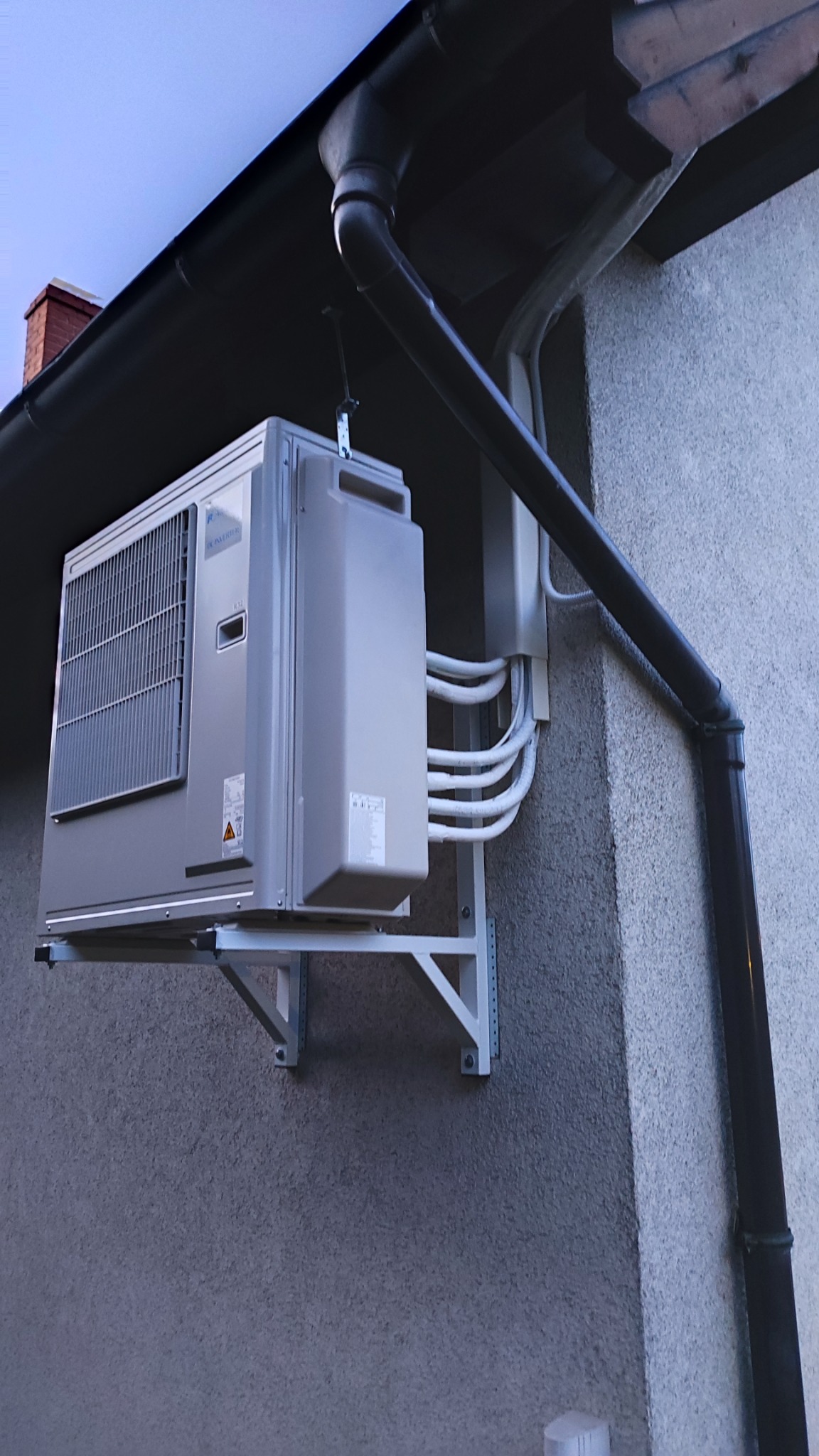 Zewnętrzna jednostka klimatyzacji zamontowana na ścianie budynku, z widocznymi przewodami i rynną spustową.