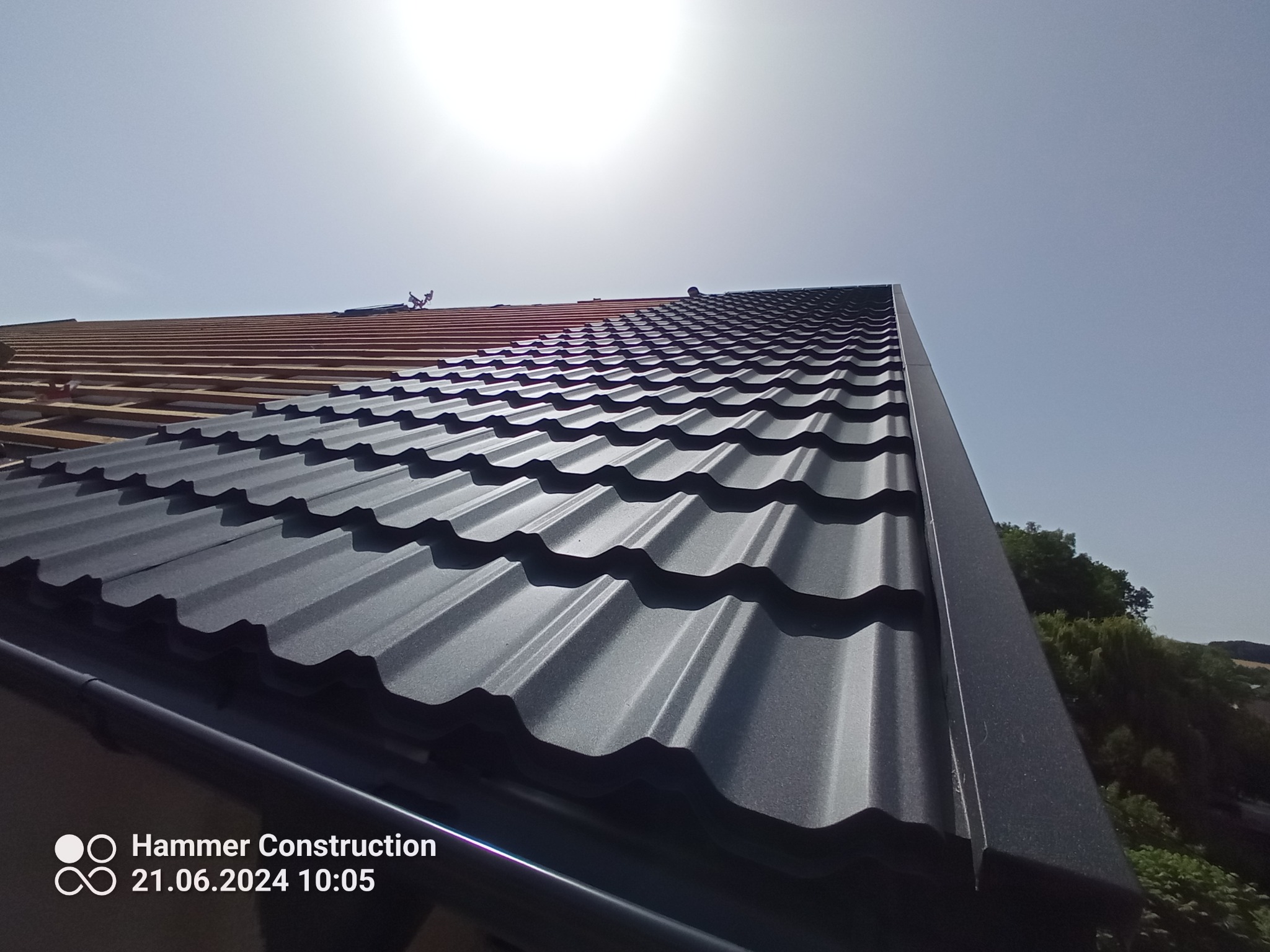 Częściowo pokryty czarną blachodachówką dach w trakcie wymiany, widoczna więźba dachowa i zamontowana rynna, ujęcie pod kątem w słoneczny dzień.