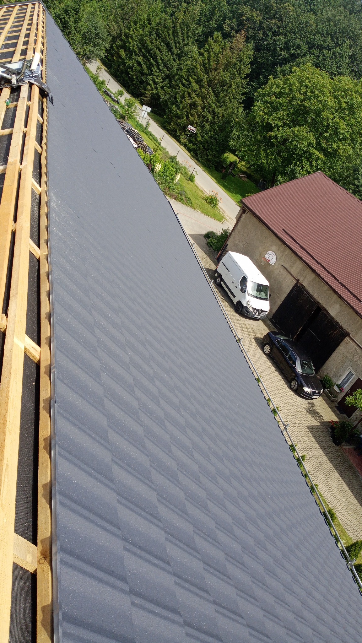 Montaż szarej blachodachówki na drewnianej konstrukcji dachu, widok z góry na budynek gospodarczy i podjazd z dwoma samochodami.