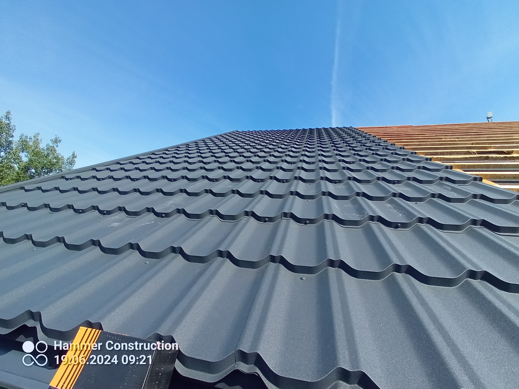 Nowe, grafitowe pokrycie dachowe z blachy trapezowej, częściowo ułożone, widoczne drewniane łaty pod spodem, błękitne niebo w tle.