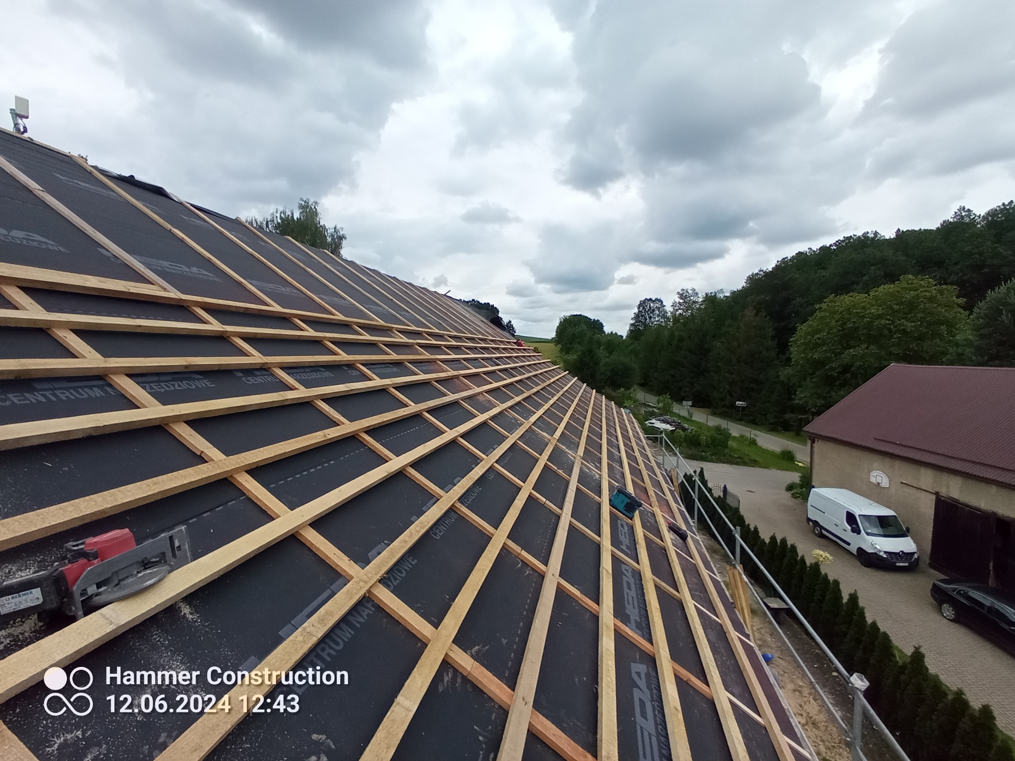 W trakcie montażu konstrukcji drewnianej pod pokrycie dachowe, widoczne deski i membrana dachowa, w tle budynek gospodarczy i samochody
