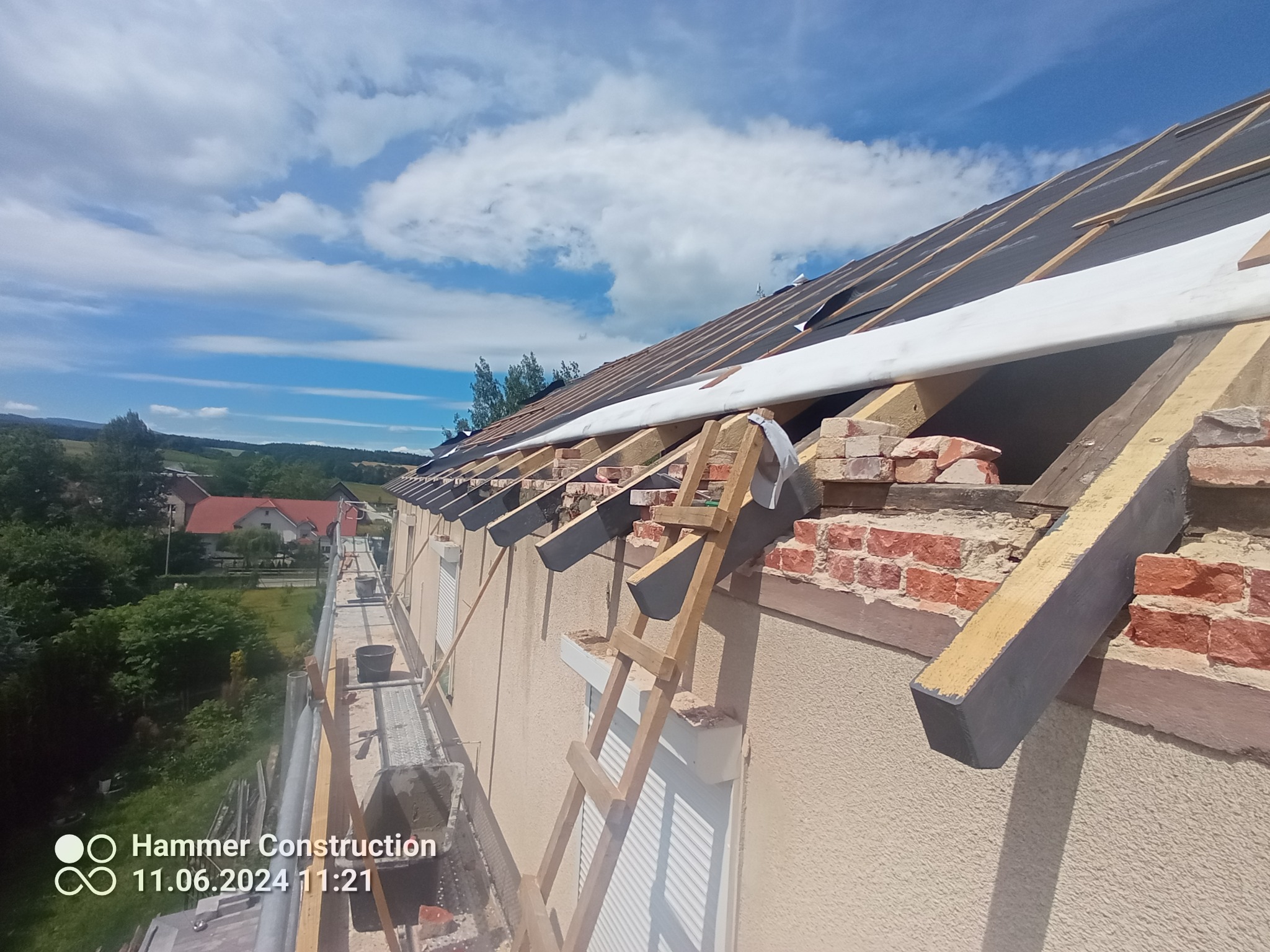 Demontaż starego dachu z ceglaną obróbką komina, widoczna więźba dachowa i elementy izolacji, rusztowanie z boku budynku, słoneczny dzień.