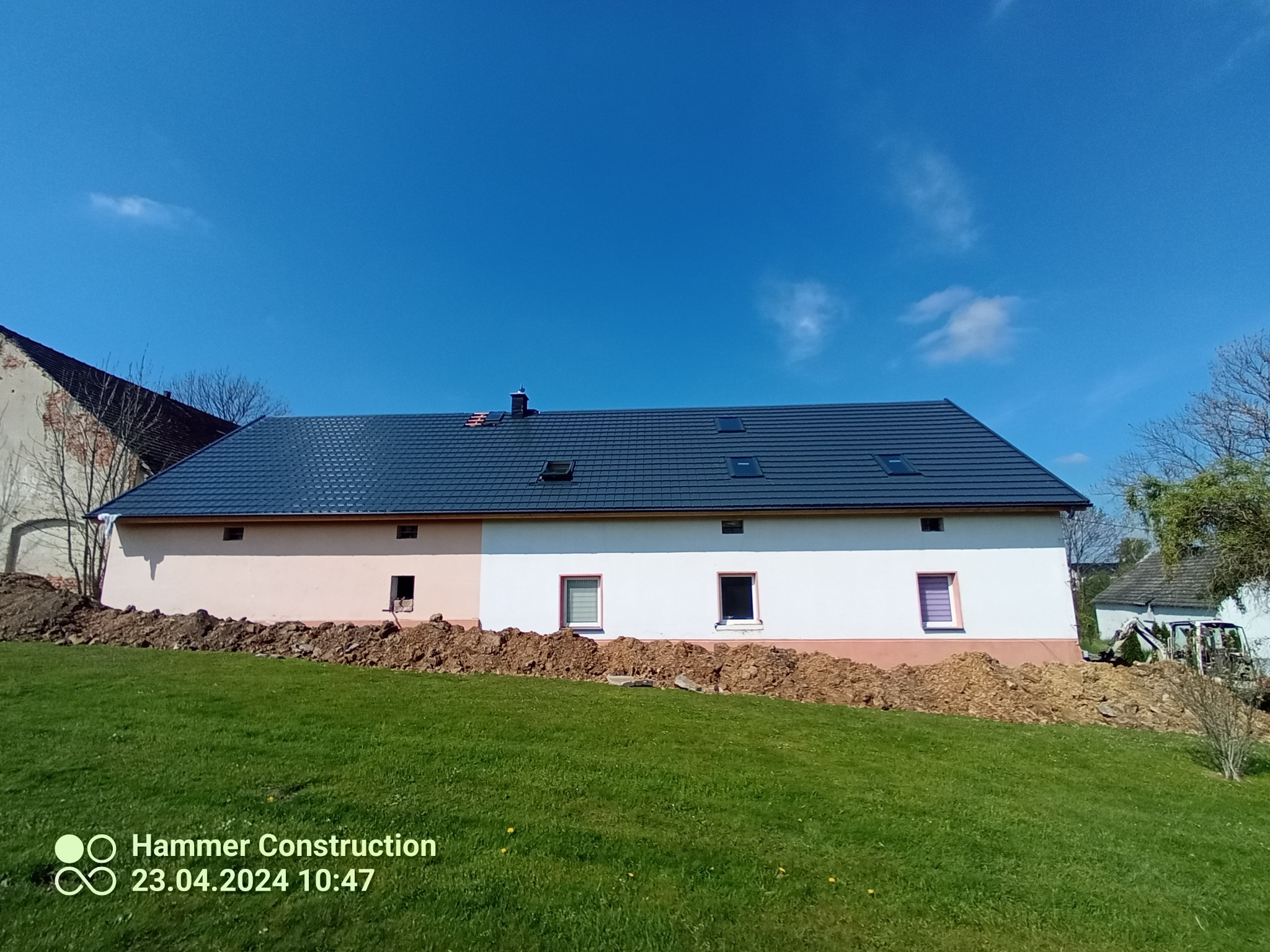 Budynek z nowym, ciemnym dachem z dachówki, widoczne okna dachowe i komin, ziemia naruszona wokół budynku, zielony trawnik, błękitne niebo.