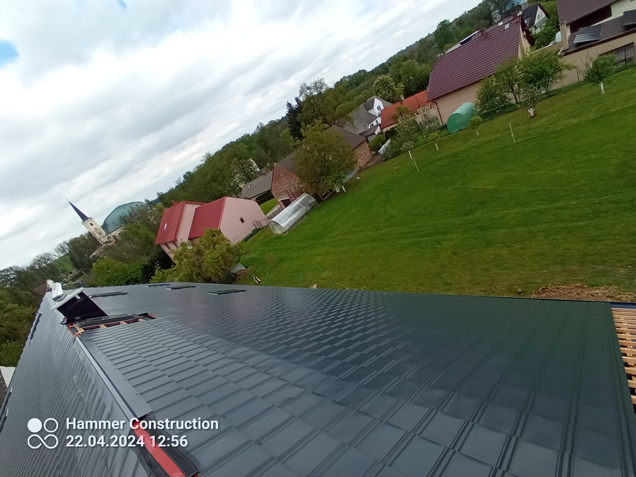 Nowy, grafitowy dach z blachy trapezowej w trakcie montażu, widok z góry na tle wiejskiego krajobrazu z kościołem i domami.