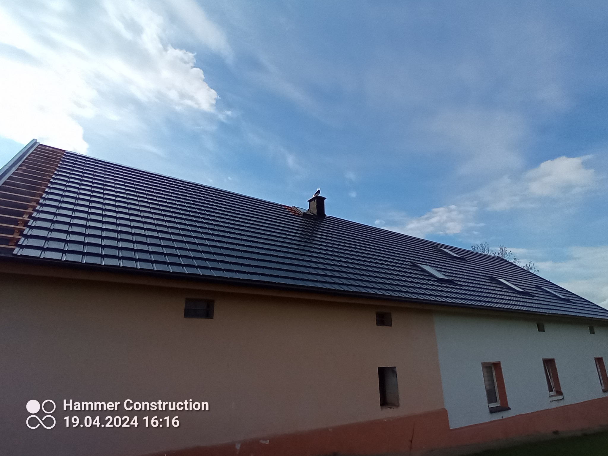 Częściowo zdemontowany dach z widoczną konstrukcją drewnianą i nową, czarną dachówką, komin z ptakiem na szczycie, budynek o dwóch odcieniach elewacji pod błękitnym niebem.