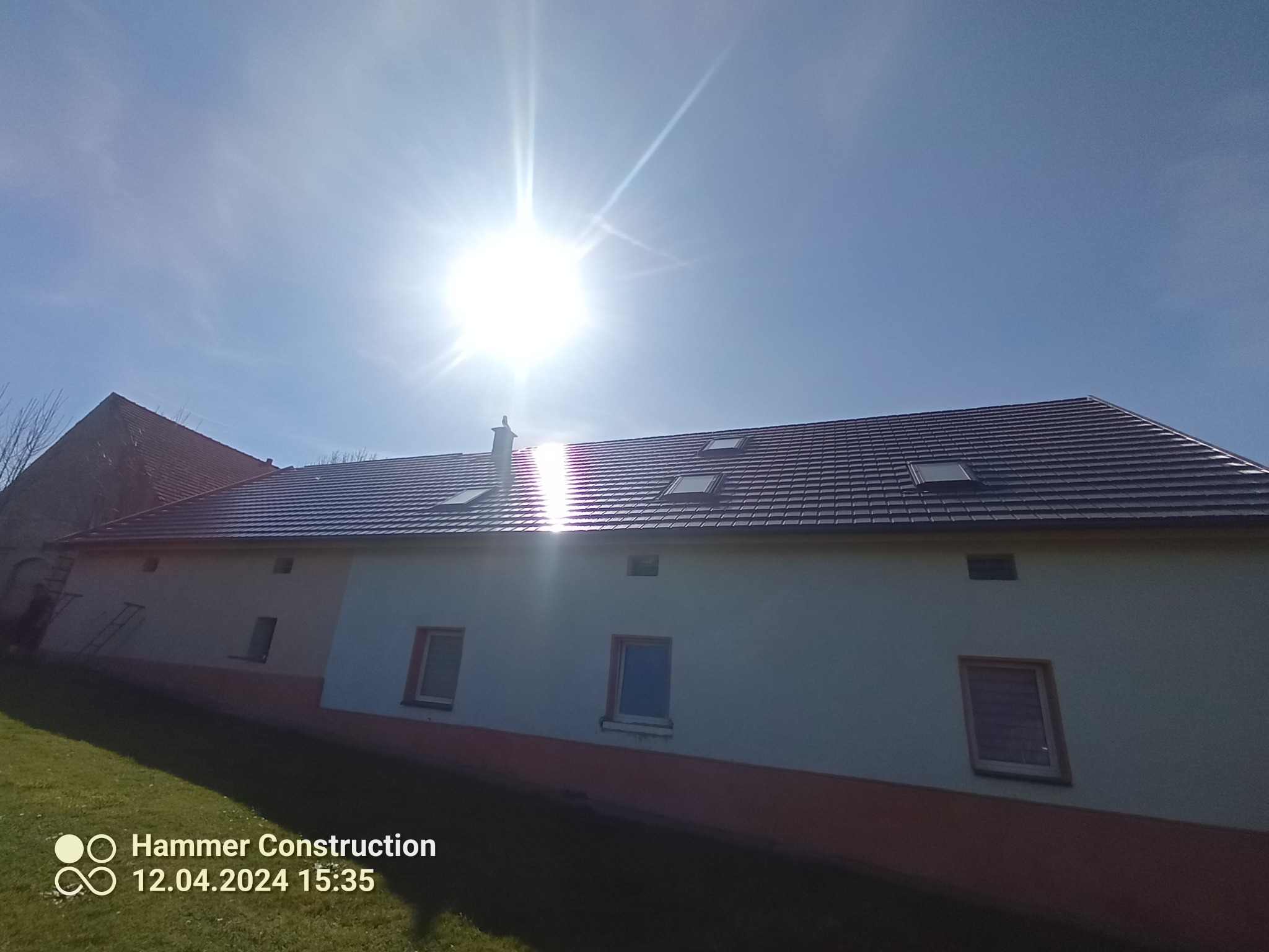 Budynek z nowym, ciemnym dachem z dachówką, widoczne okna dachowe, jasne słońce na błękitnym niebie.