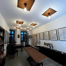 Jasne wnętrze gabinetu z brązowym kozetką, biurkiem, regałem z książkami, dekoracyjnym drzewkiem i mapami anatomicznymi na ścianie, oświetlone kwadratowymi lampami sufitowymi.