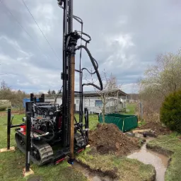 Mobilna wiertnica na gąsienicach, przygotowana do wiercenia, obok wykopany rów wypełniony wodą, zbiornik wody, pochmurne niebo w tle.